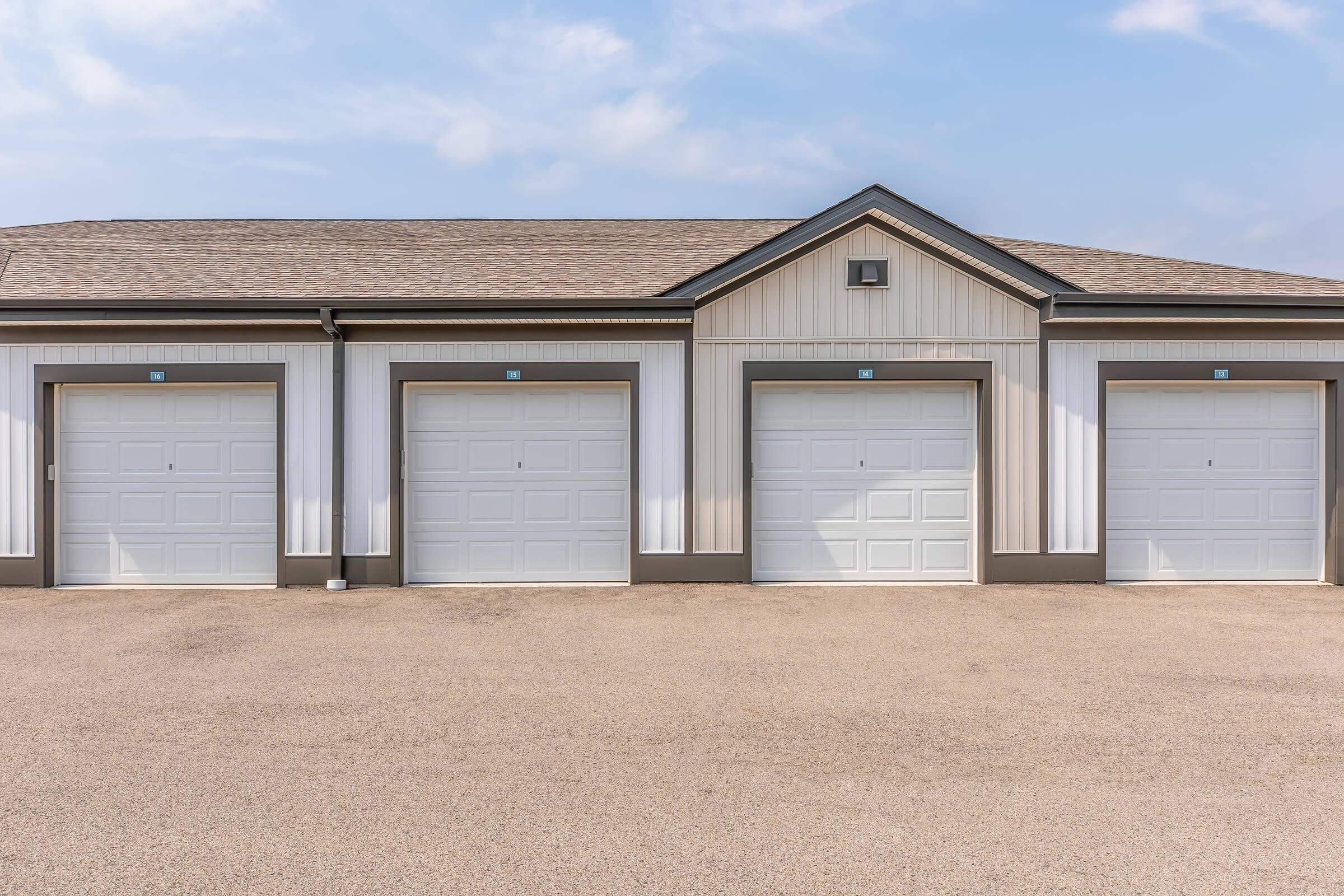 Three closed garage doors in a row on a light-colored building with a sloped roof. The facade features a mix of siding styles, and a clear blue sky is visible above. The ground is paved and slightly textured. The overall setting appears clean and well-maintained.