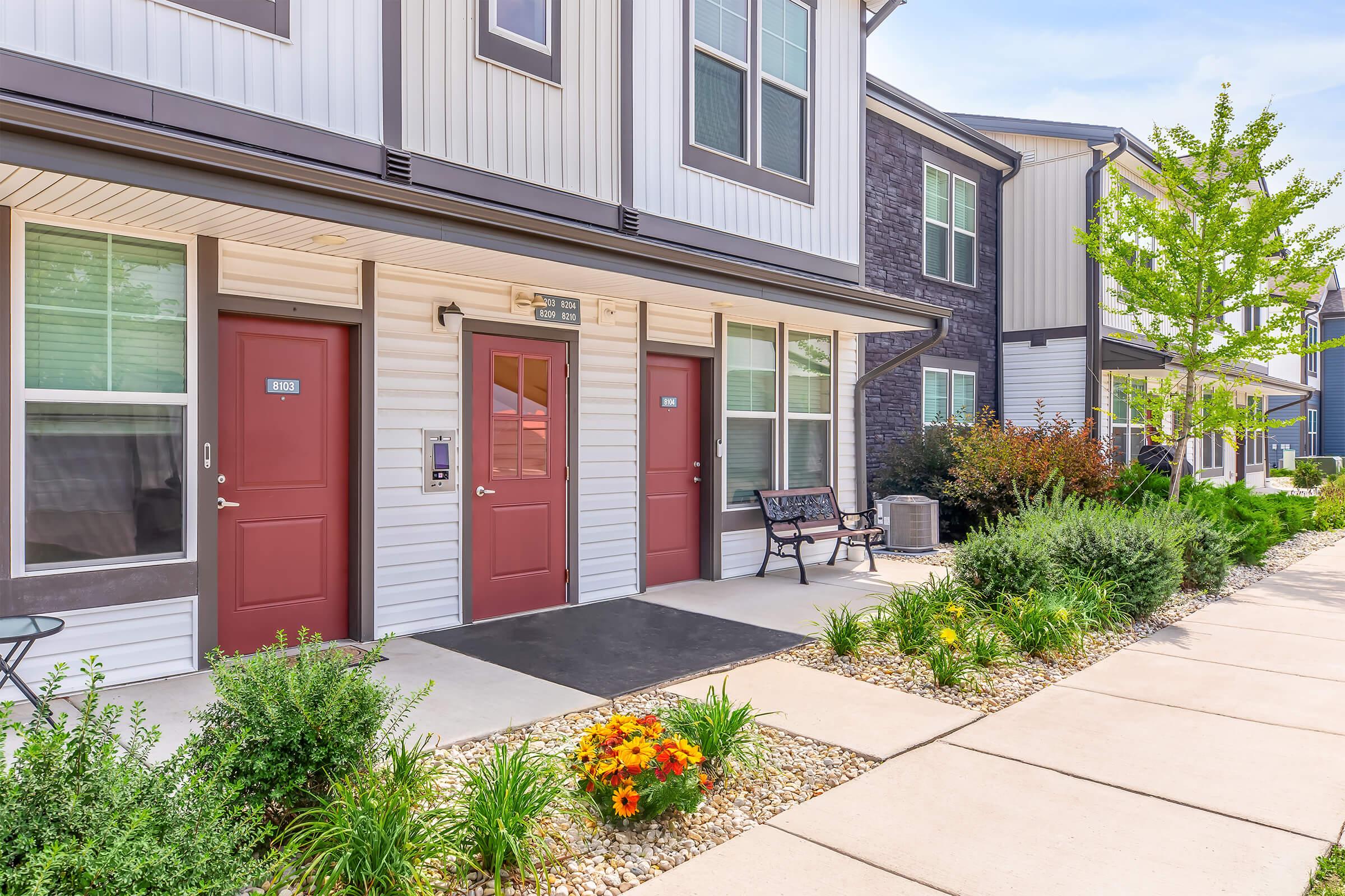 Modern apartment complex exterior featuring two entry doors with red accents, a landscaped pathway with flower beds, and benches. The surrounding greenery includes shrubs and small trees, adding to the inviting atmosphere of the residential area. Bright, sunny day with clear skies.