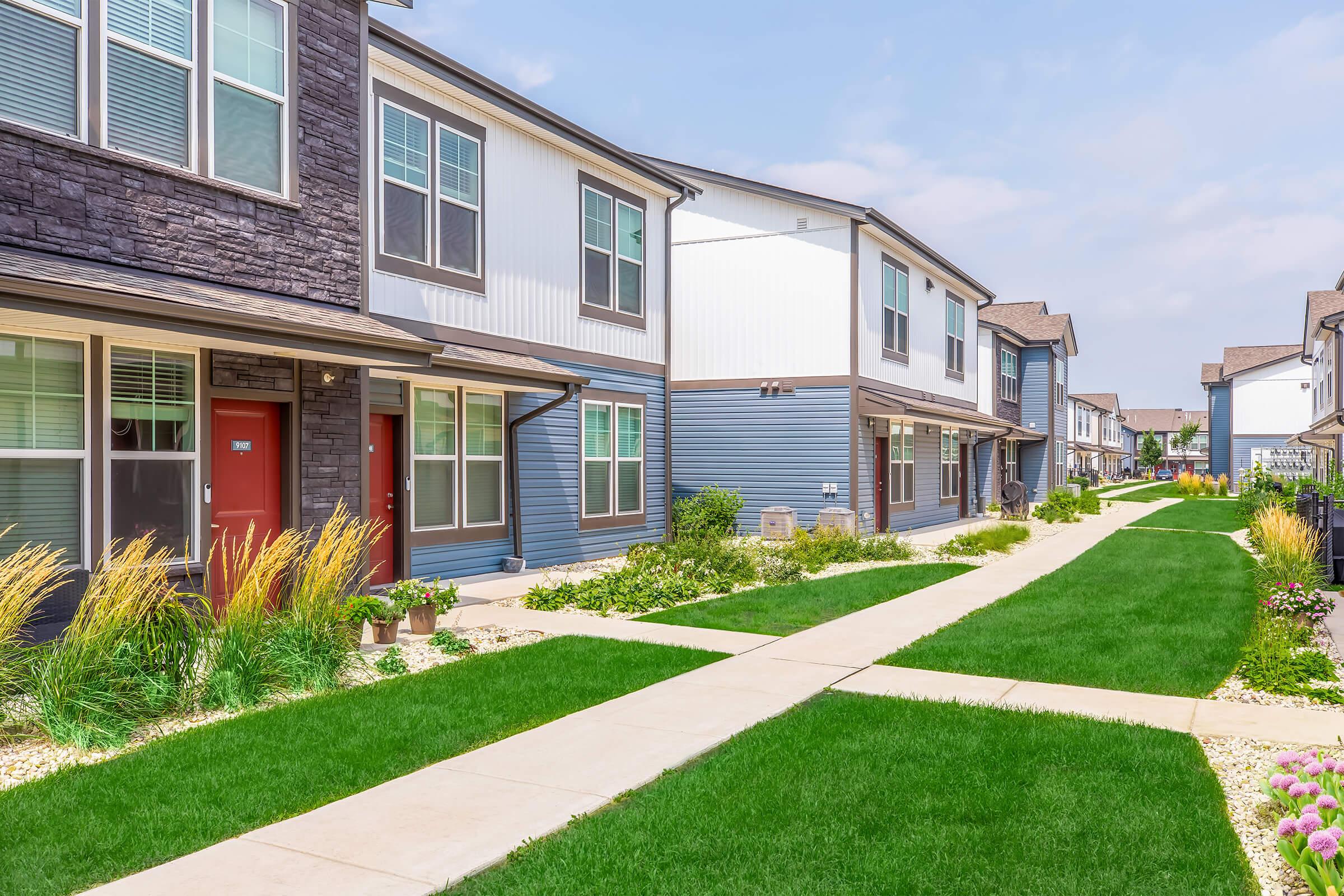 A view of a landscaped pathway between two-story residential buildings. The pathway is bordered by well-maintained grass and flower beds, with modern architecture featuring a mix of siding materials. Blue sky and soft clouds complete the pleasant suburban scene.