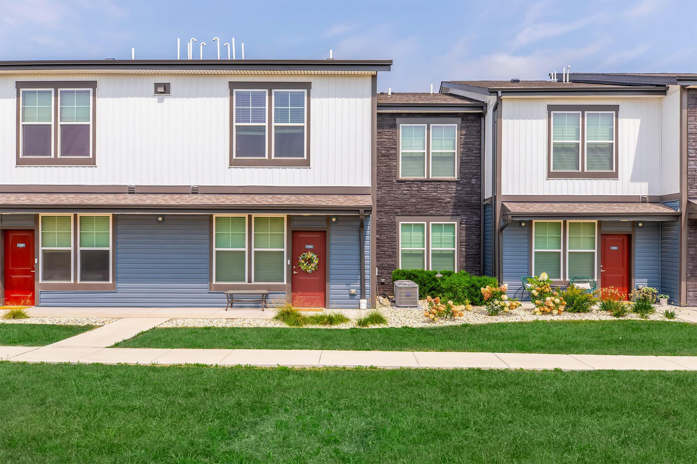 Two modern townhouses side by side, featuring light blue and dark brown exteriors. Each unit has a front porch with a bench, red doors, and large windows. A small garden with flowers and shrubs is visible in front of the houses, with a well-maintained grassy area in the foreground.