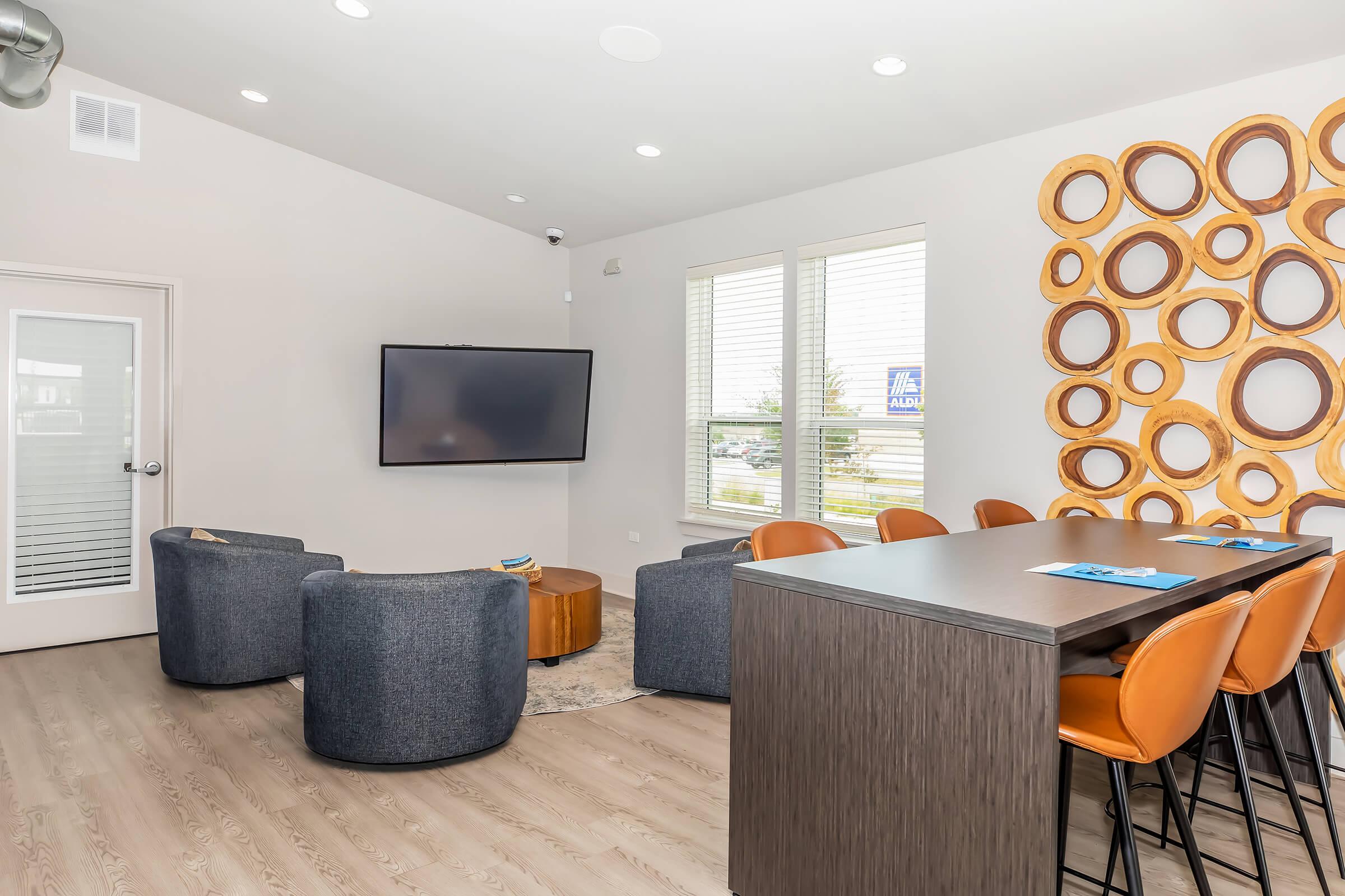 A modern lounge area featuring a large television mounted on a wall, surrounded by light-colored walls and large windows. Plush blue chairs and a round wooden coffee table are arranged for seating, alongside a sleek bar with stylish orange stools. Decorative circular mirrors adorn the wall.