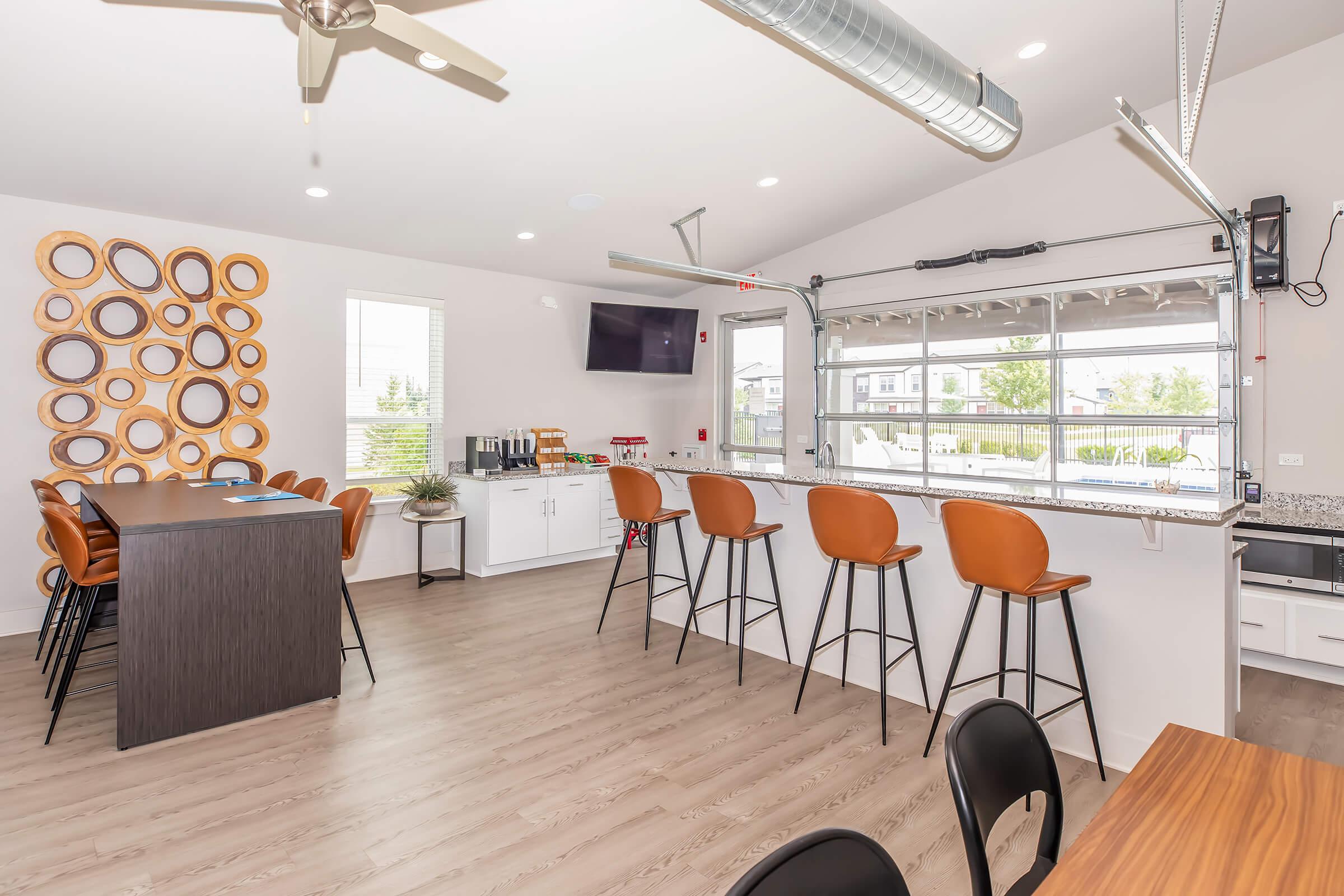 A modern kitchen and dining space featuring a wooden bar with high stools, a marble countertop, and a wall adorned with circular wall decor. Large windows provide natural light, and there is a TV mounted on the wall. The space is designed for casual dining and social gatherings.
