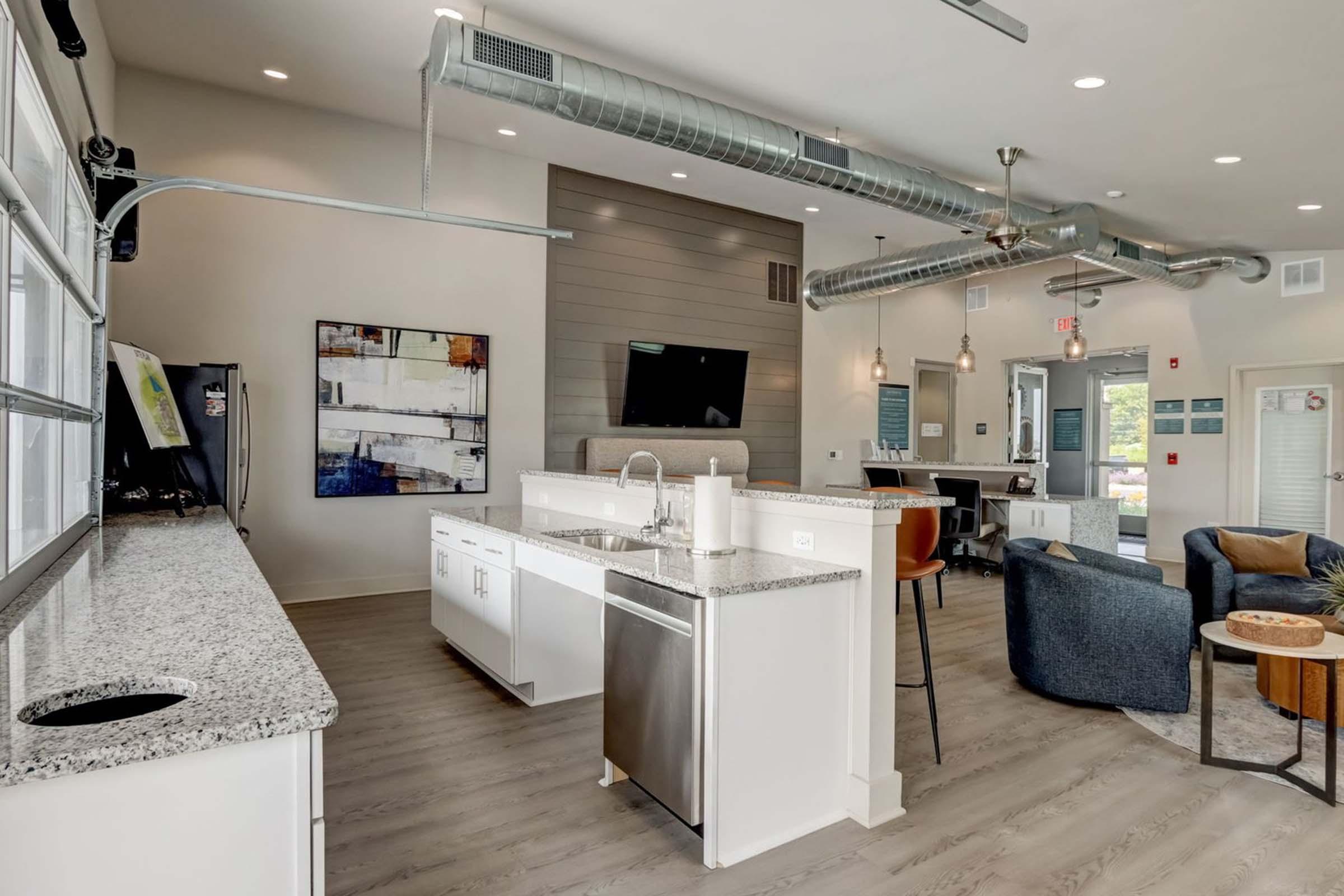 Modern kitchen and living area featuring a granite countertop, stainless steel appliances, and an open layout. The space has large windows, contemporary furniture, and decorative artwork on the wall. Exposed ductwork and sleek design elements enhance the stylish ambiance.
