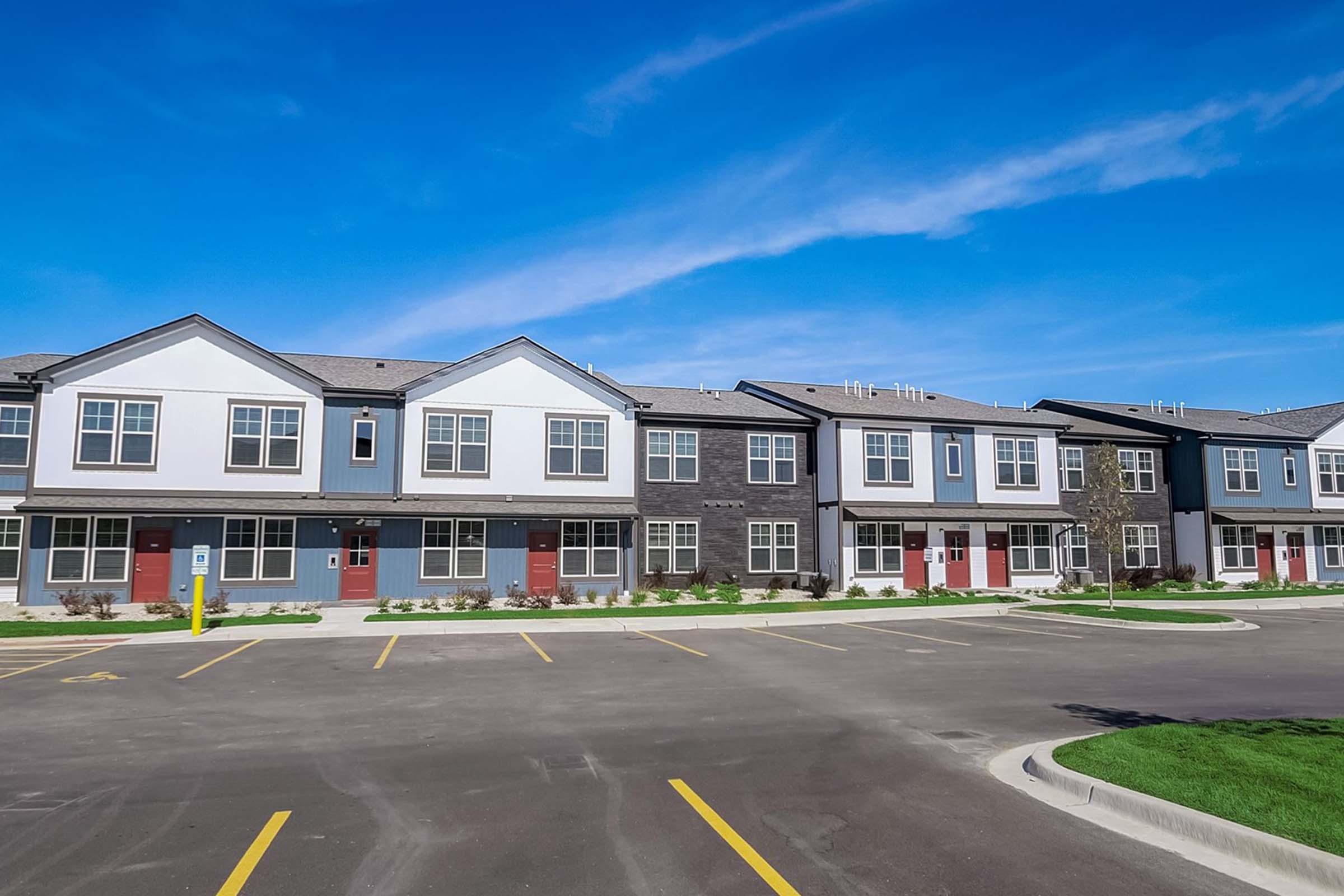 A modern residential building featuring multiple two-story units with a combination of gray and white exteriors. The complex includes a paved parking area and well-maintained landscaping under a clear blue sky.
