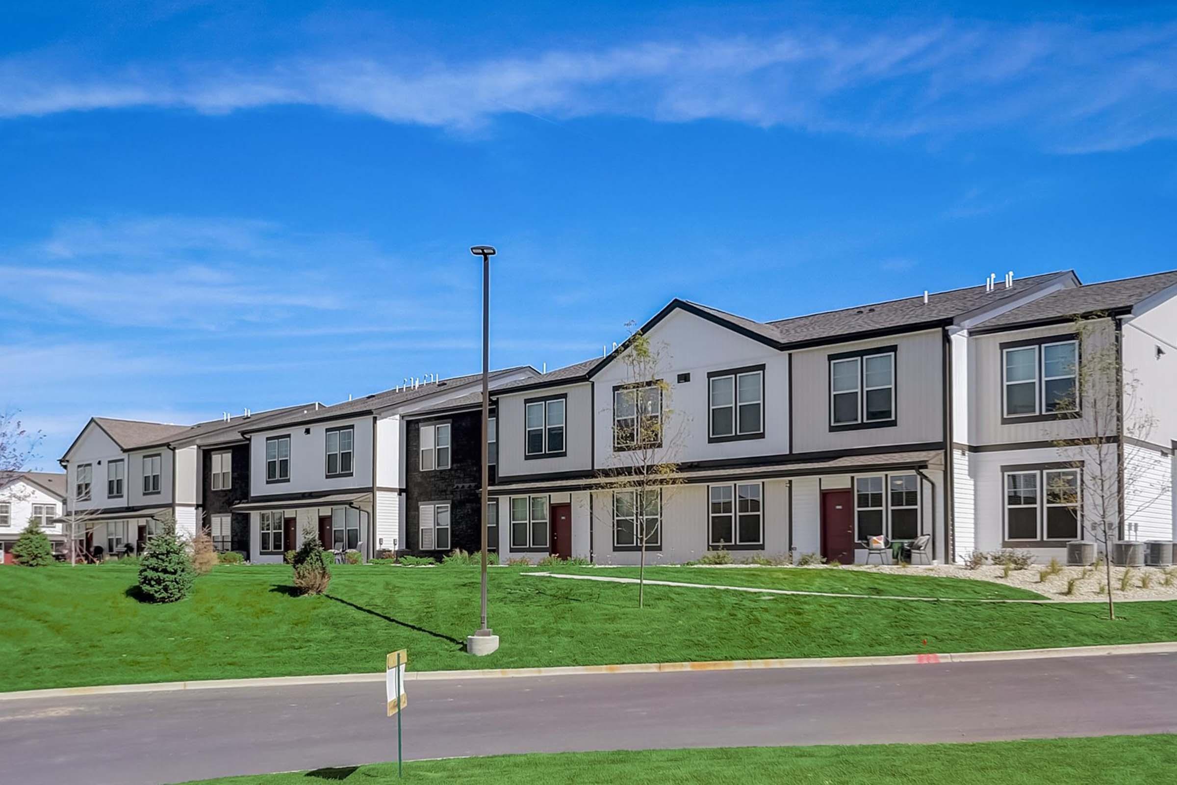 A row of modern residential buildings with multiple units, featuring a mix of colors and architectural styles. Each unit has large windows and colorful front doors, situated on a well-maintained lawn under a clear blue sky. A paved pathway runs in front of the buildings.