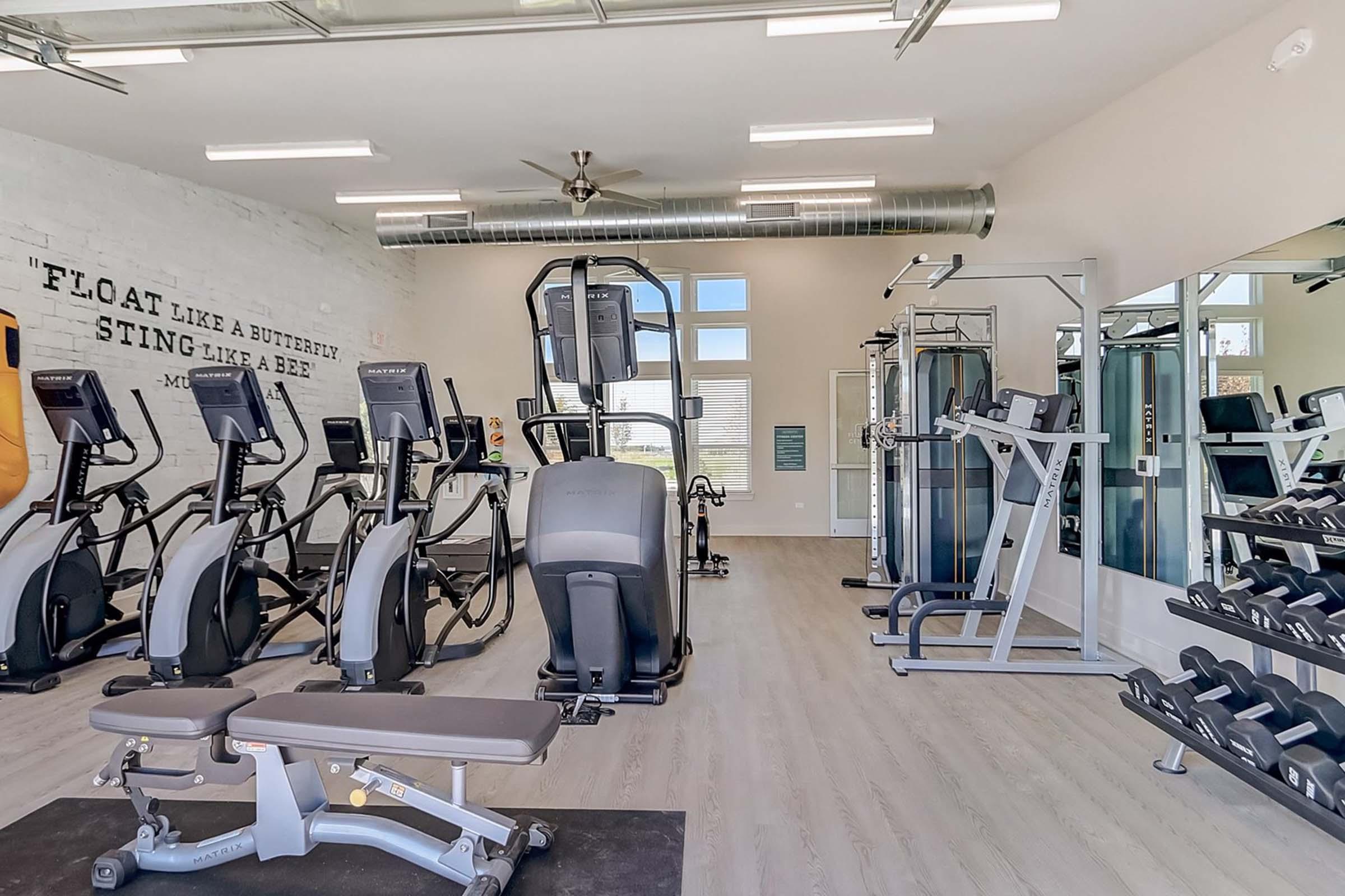 A modern gym interior featuring cardio machines, weight training equipment, a bench press, and mirrors. The walls are adorned with inspirational quotes about fitness, and large windows allow natural light to brighten the space.