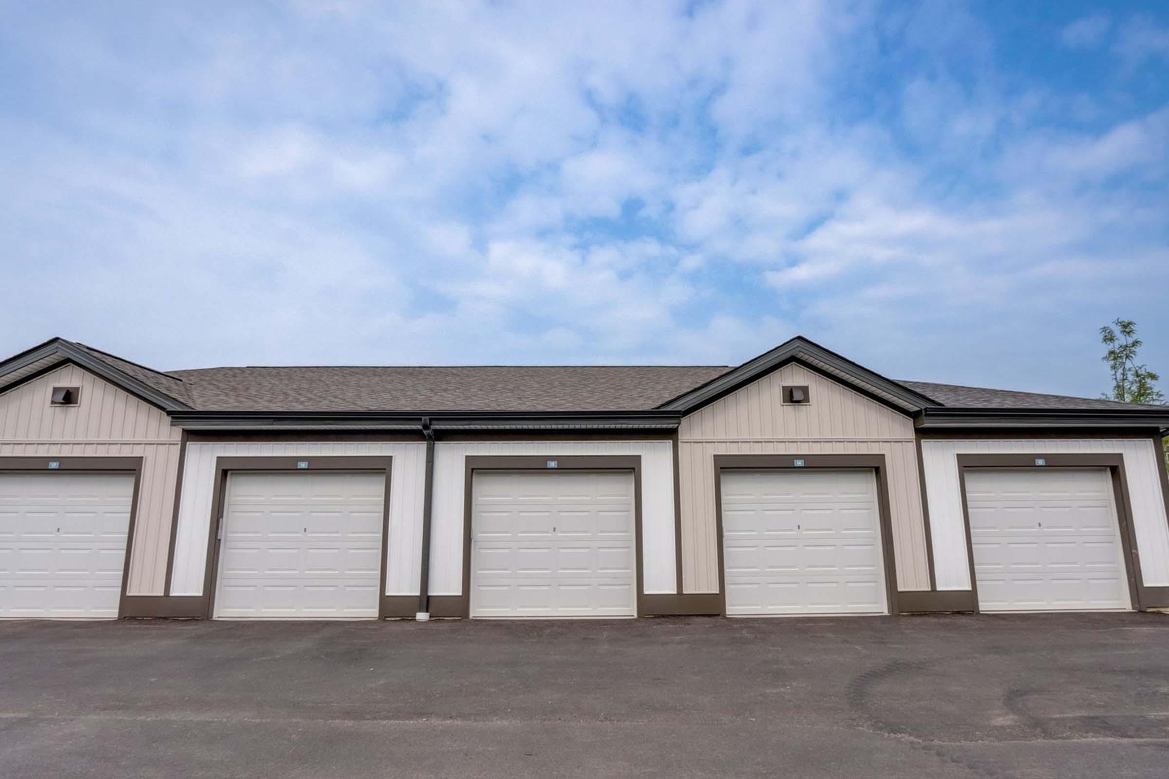 A row of six identical storage garages with white doors, set against a blue sky with clouds. The garages have a simple, modern design with gray trim, and are located on a paved surface.