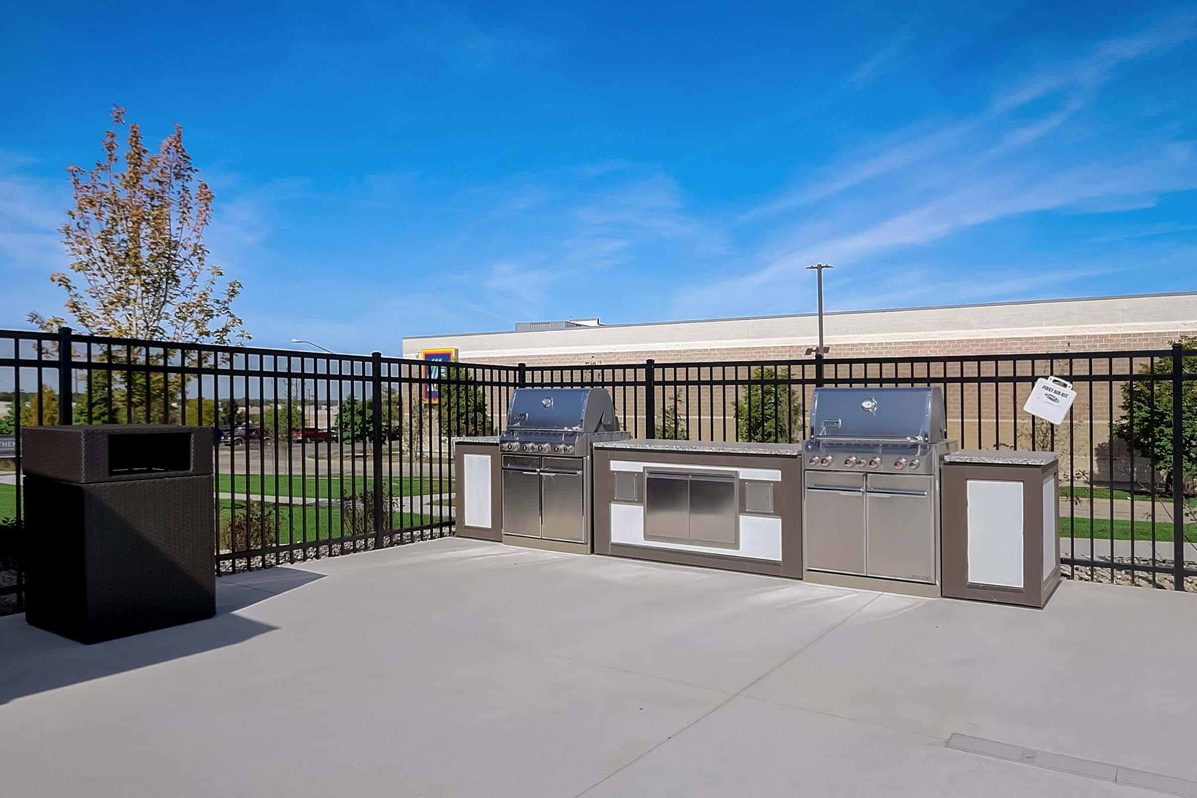 Outdoor grilling area featuring two built-in grills, a large counter space, and a trash bin nearby. The setting is enclosed by a black fence, with a clear blue sky and trees in the background. Ideal for outdoor cooking and gatherings.
