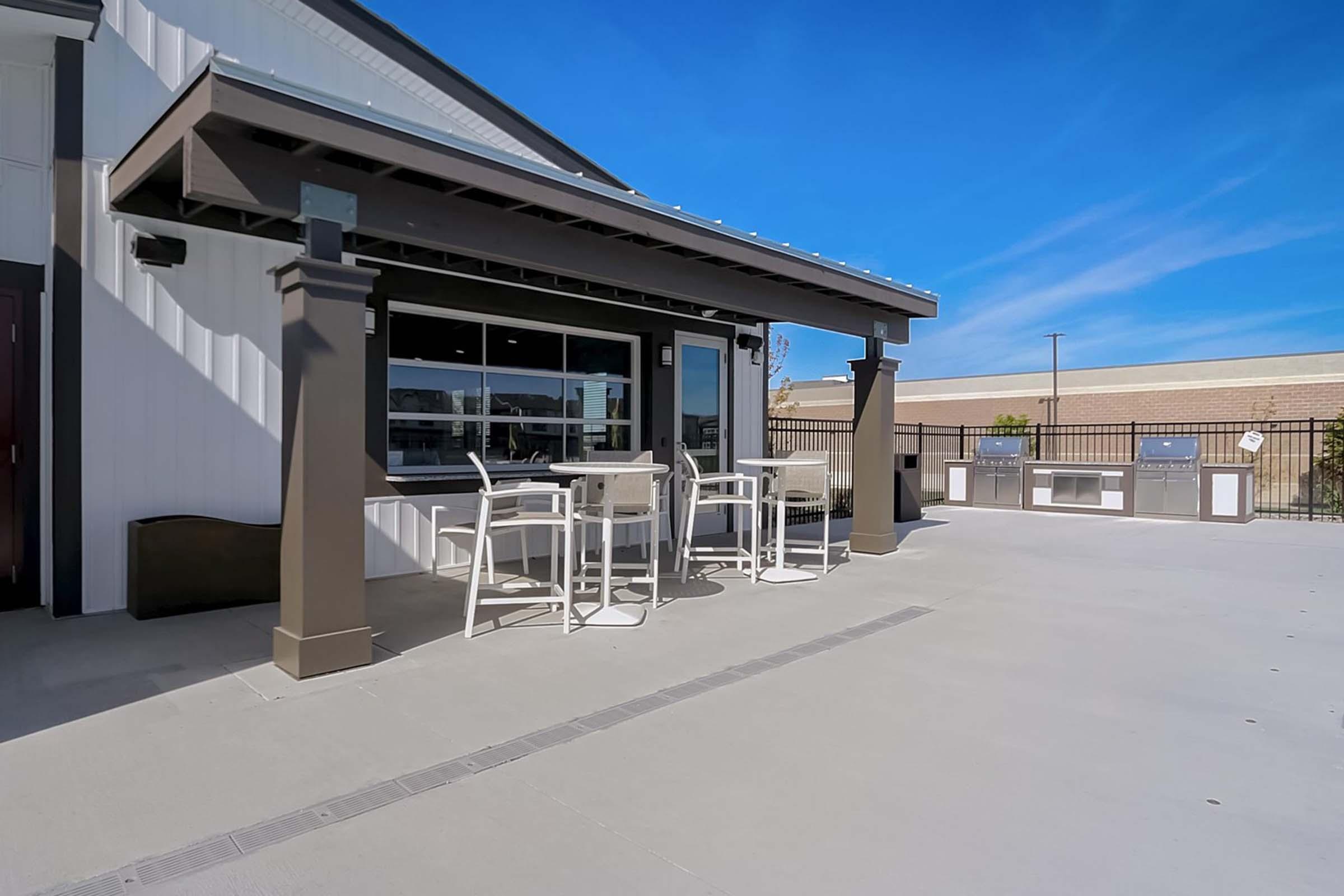 Outdoor patio area with modern seating, featuring a table and chairs under a shaded structure. The backdrop includes a clear blue sky and a fence, providing a relaxing space for gatherings or dining outside.
