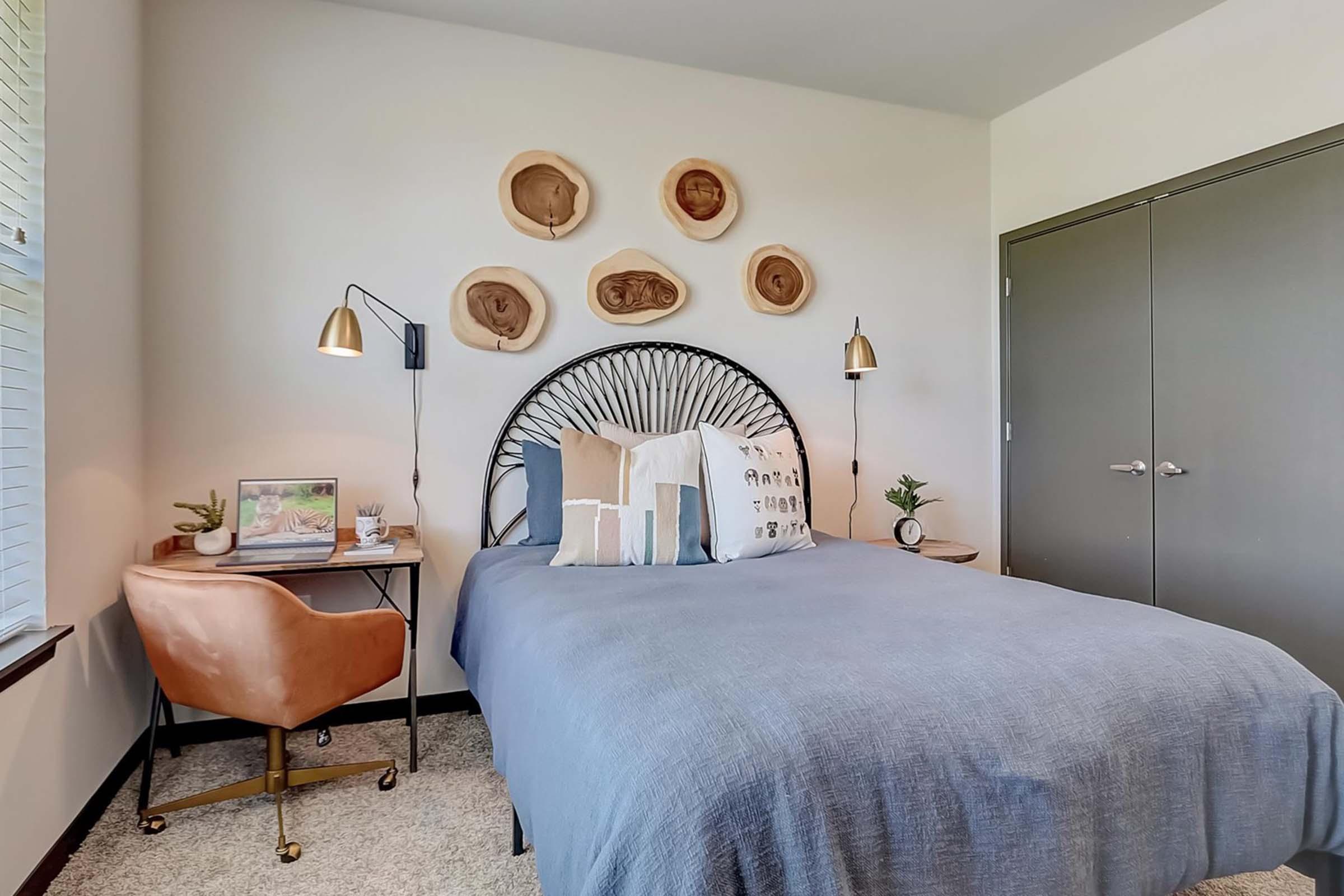 A cozy bedroom featuring a bed with a blue bedspread and decorative pillows. Above the bed are wooden wall art pieces. To the side, there is a small desk with a chair and a plant, along with stylish lighting fixtures. The overall decor is modern and inviting, with neutral tones and soft textures.