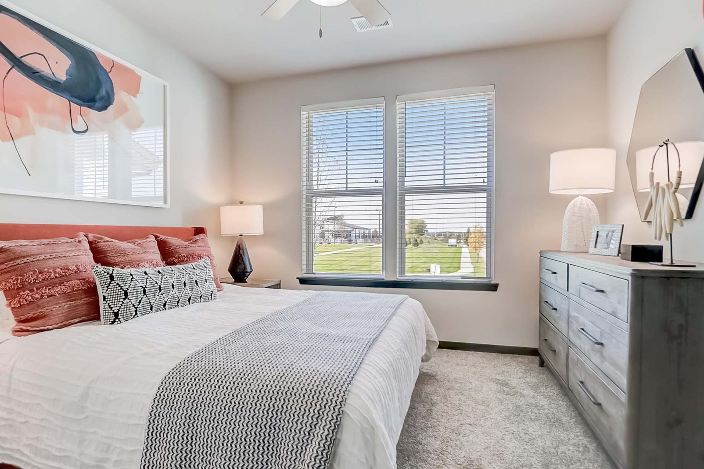A cozy bedroom featuring a white bed spread adorned with decorative pillows, a nightstand with a lamp, and a dresser. Two large windows provide natural light and a view of grassy outdoors. A colorful abstract painting hangs above the bed, adding a modern touch to the space.