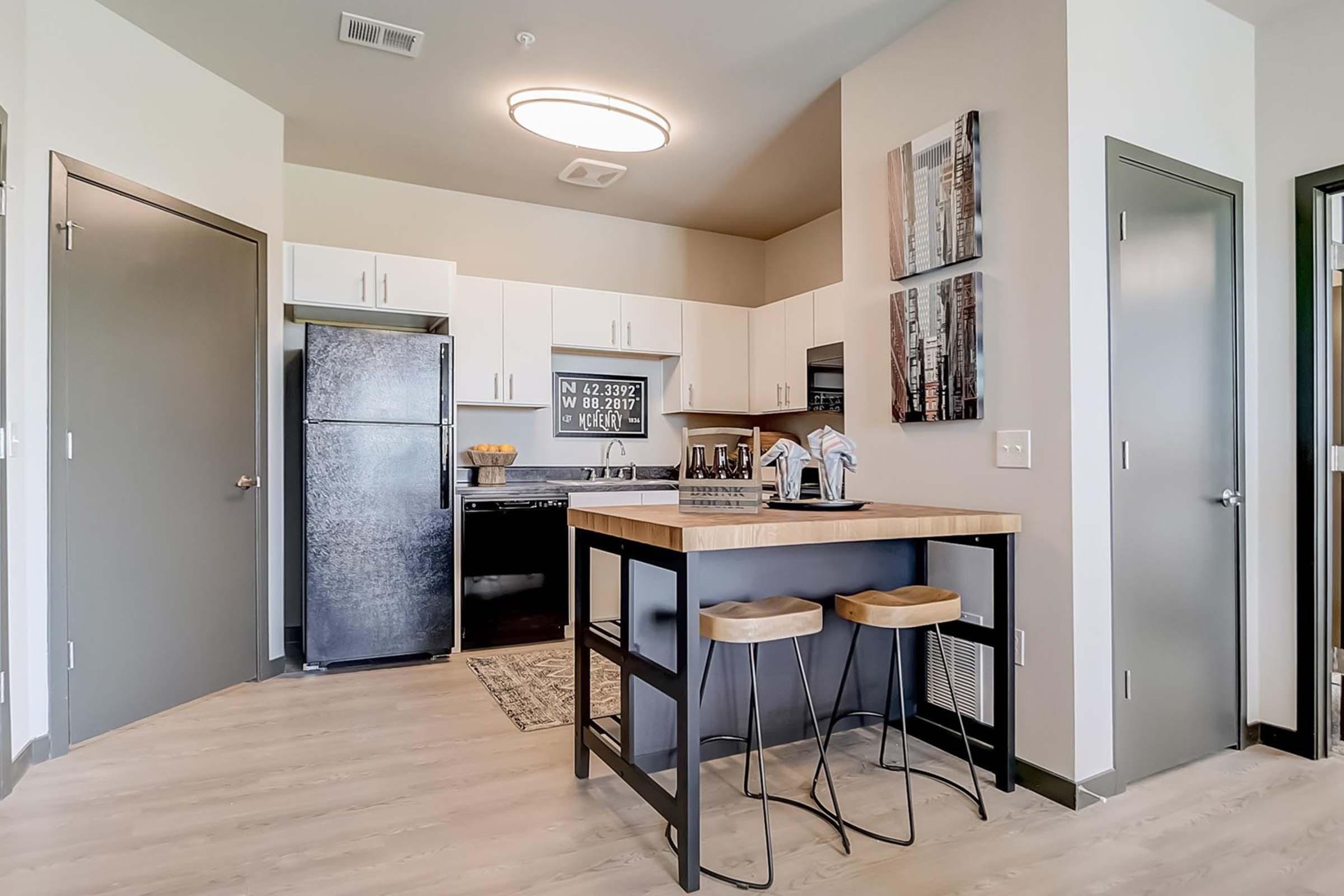 Modern kitchen in an apartment featuring a small dining area with two stools, a wooden countertop, a black refrigerator, and white cabinetry. Wall art is displayed adjacent to the kitchen area, and the flooring is light-colored wood. The space is well-lit and stylishly decorated.