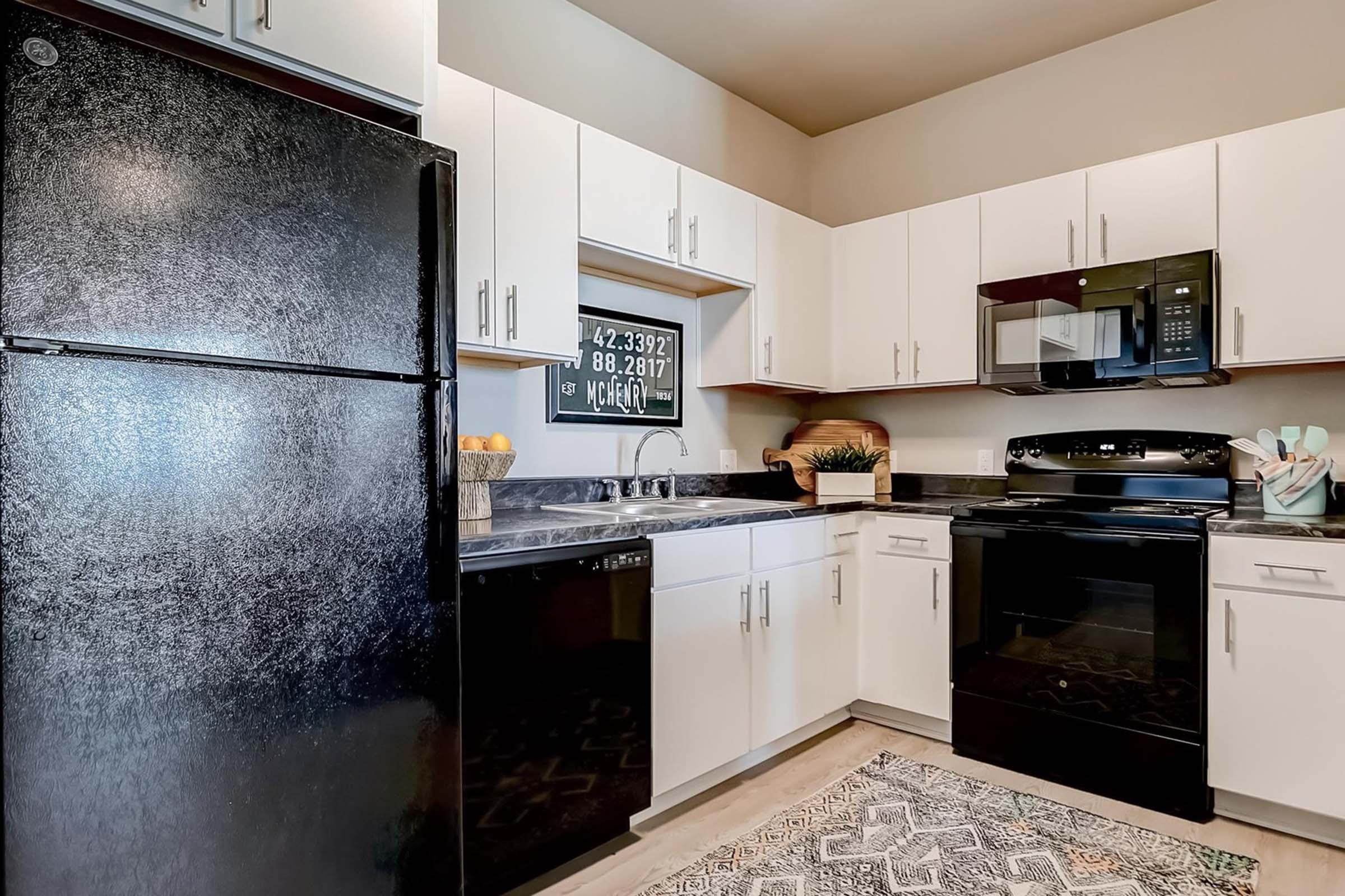 A modern kitchen featuring white cabinetry, black appliances including a refrigerator, oven, and microwave. A decorative bowl of fruit is on the countertop, and a patterned rug lies on the floor. A window with a framed sign is visible, adding to the room's aesthetic.