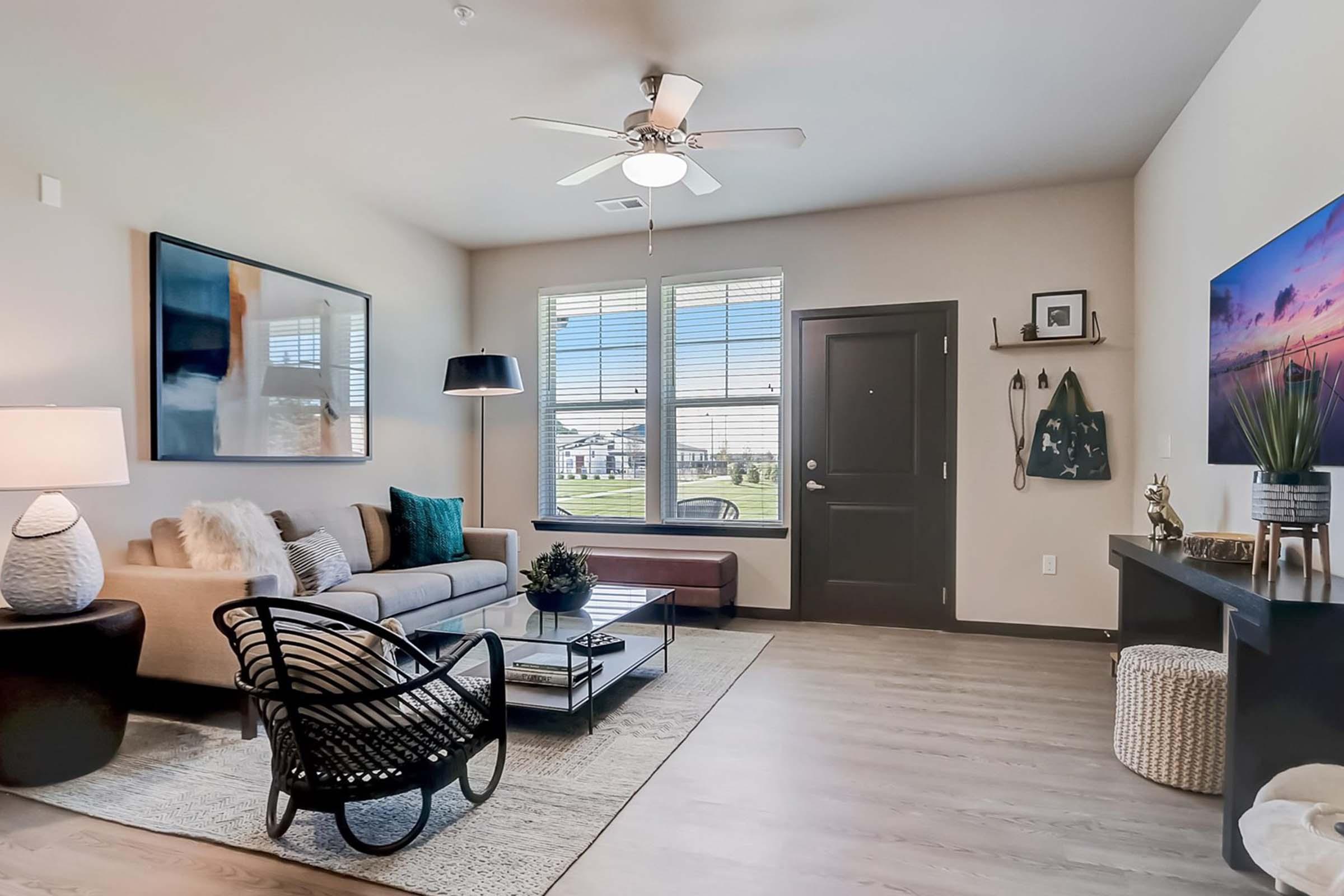 A modern living room featuring a beige sofa with decorative pillows, a glass coffee table, and a black wicker chair. There’s a ceiling fan, large windows with blinds allowing natural light, and a sleek entrance door. Wall art and decorative items enhance the stylish ambiance.