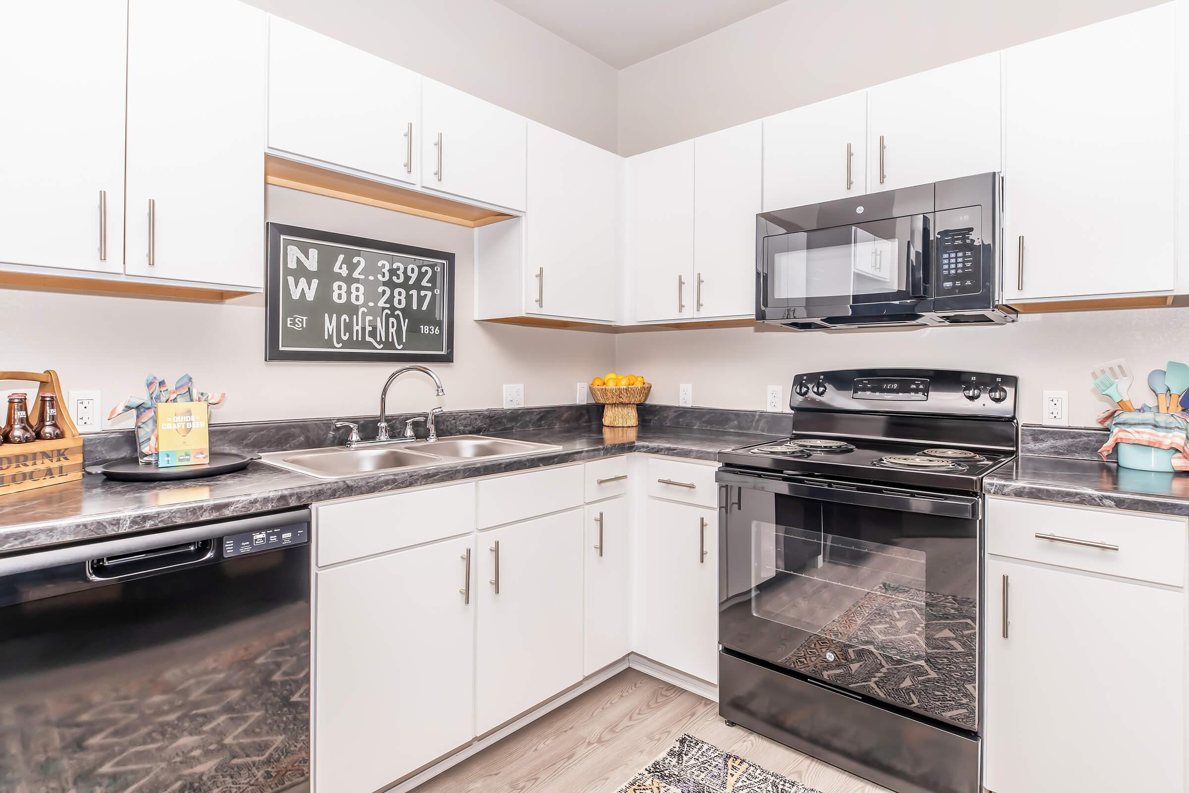 Modern kitchen featuring white cabinetry, a black countertop, a stainless steel sink with a dishwasher, a black oven, and a microwave above it. Wall decor includes a framed sign with coordinates. The space is well-lit, showcasing a cozy atmosphere with kitchen essentials and decorative items.