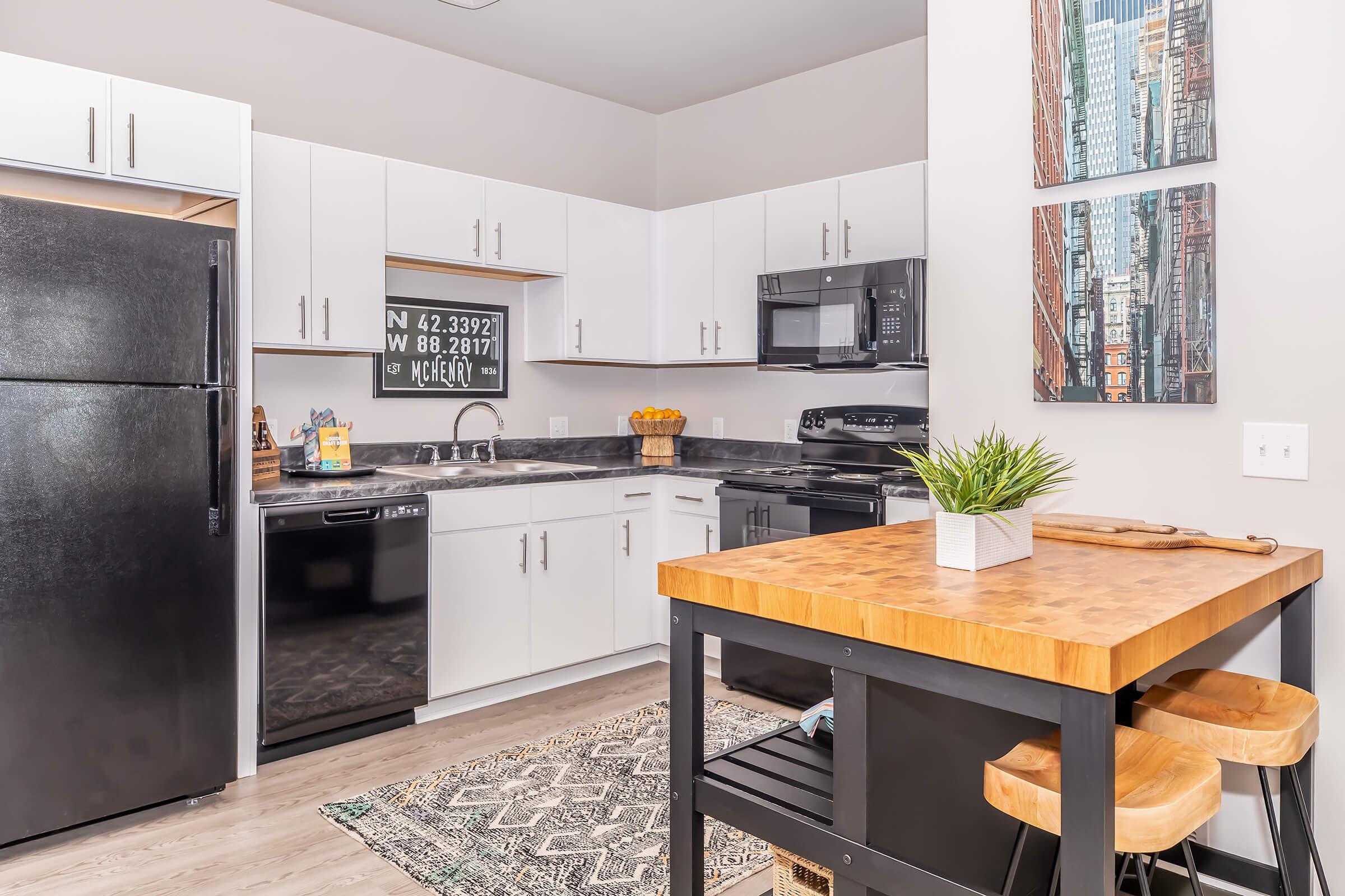 Modern kitchen featuring black appliances, white cabinets, and a wooden countertop. A small dining area with two stools is visible, along with a decorative plant. Wall art depicting cityscapes adds a stylish touch. The space is well-lit and organized, showcasing a functional layout.