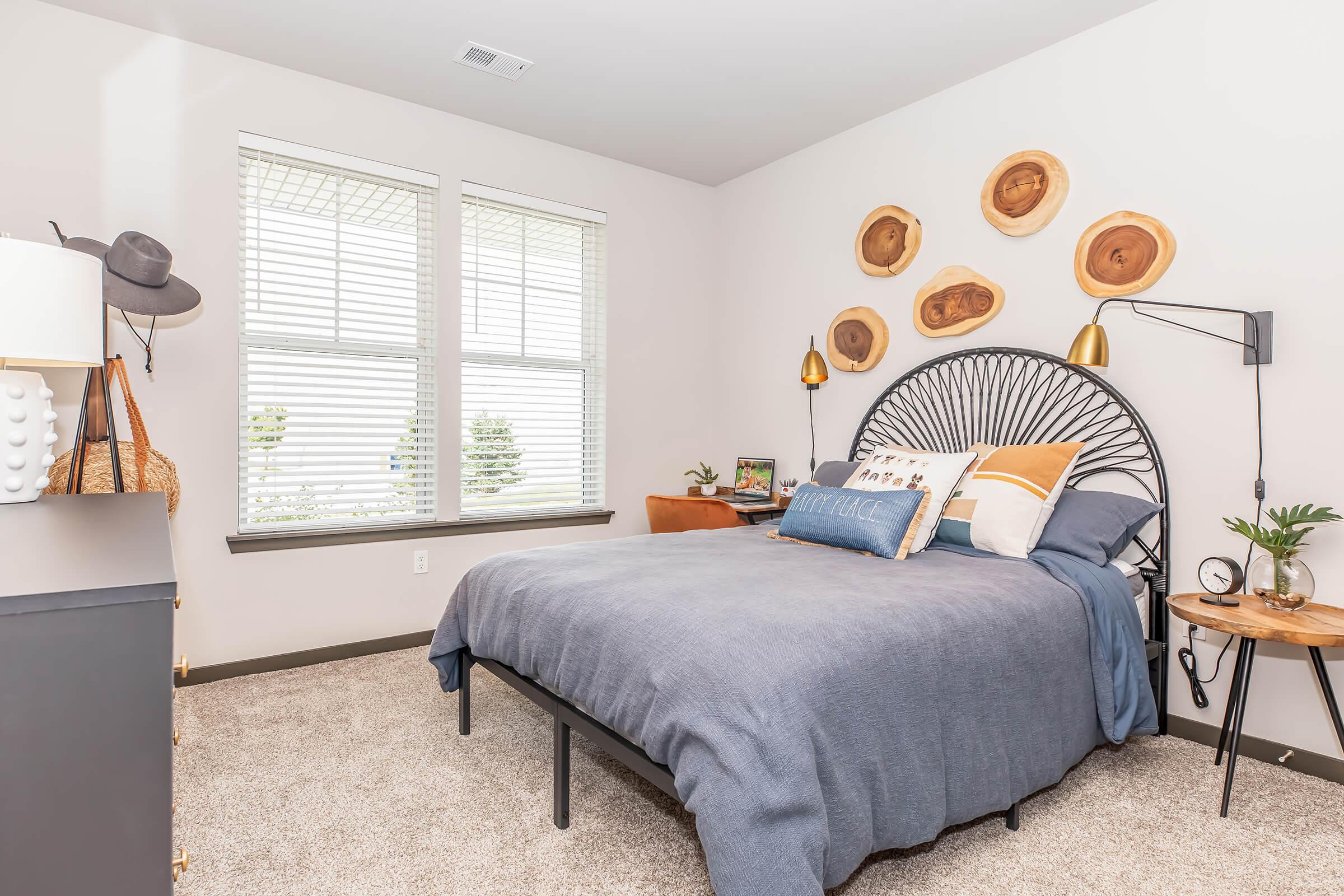 A cozy bedroom featuring a black metal bed frame with blue bedding and decorative pillows. The walls are adorned with circular wooden accents, and there are two stylish wall lamps. A small side table holds a plant, and a dark dresser is visible on the left. Large windows let in natural light.