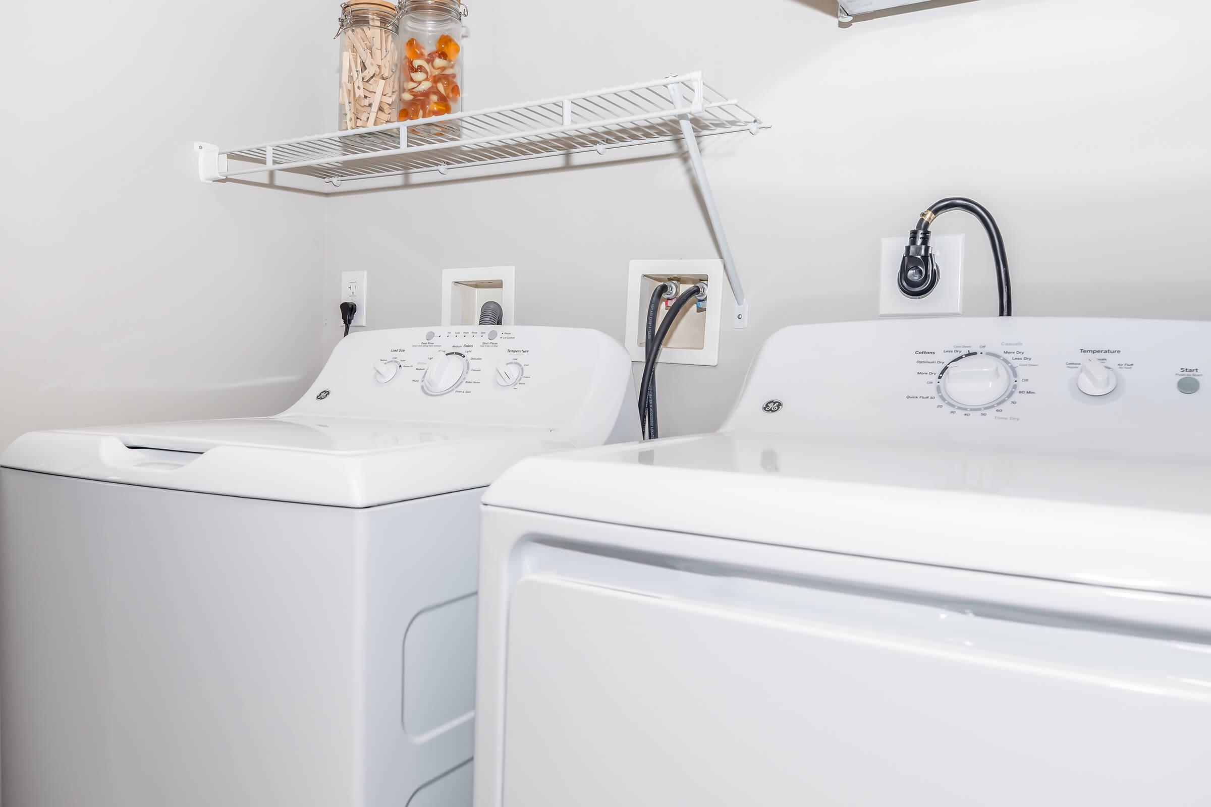 A clean laundry room featuring a white washing machine and a dryer side by side. Above, there is a shelf holding a jar with various items. The walls are painted a light color, and the machines have visible control dials and connections. The overall appearance is tidy and organized.