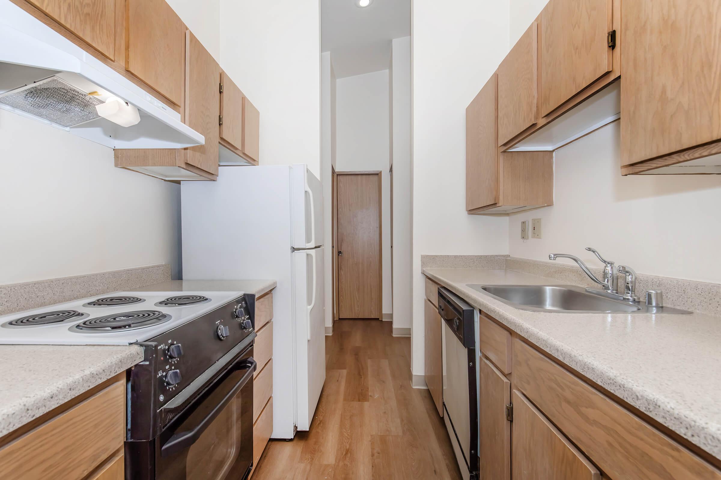 A modern kitchen featuring wooden cabinets, a white refrigerator, and a stainless steel sink. The kitchen includes a stove with a vent hood and a dishwasher. Light-colored countertops and wooden flooring enhance the bright, spacious feel. A doorway leads to another area of the home.