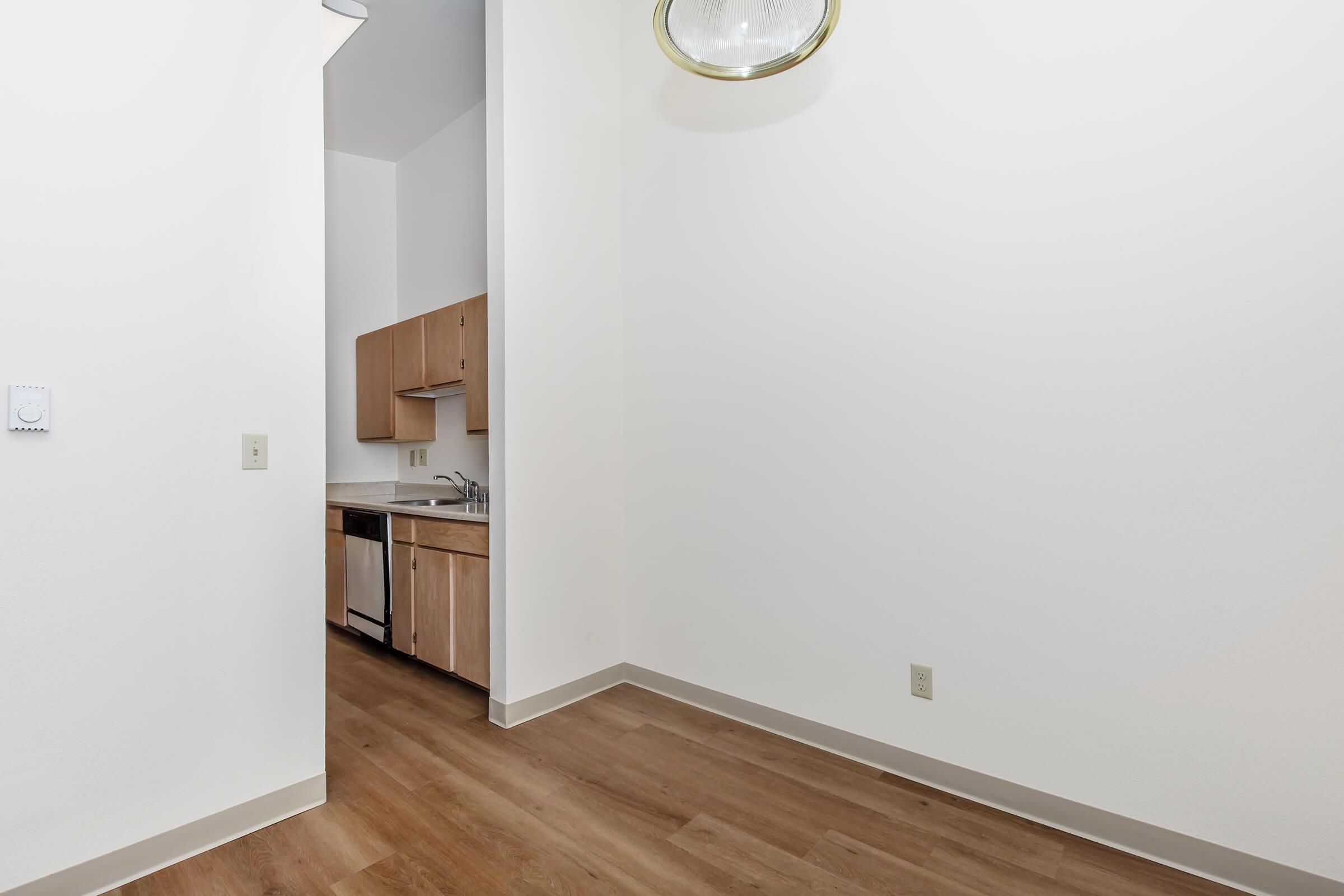 A spacious, empty room with white walls and wood flooring. There’s a small kitchenette featuring wooden cabinets and stainless steel appliances, including a refrigerator. A light fixture hangs from the ceiling, and the overall atmosphere is bright and clean, suggesting a ready-to-move-in space.