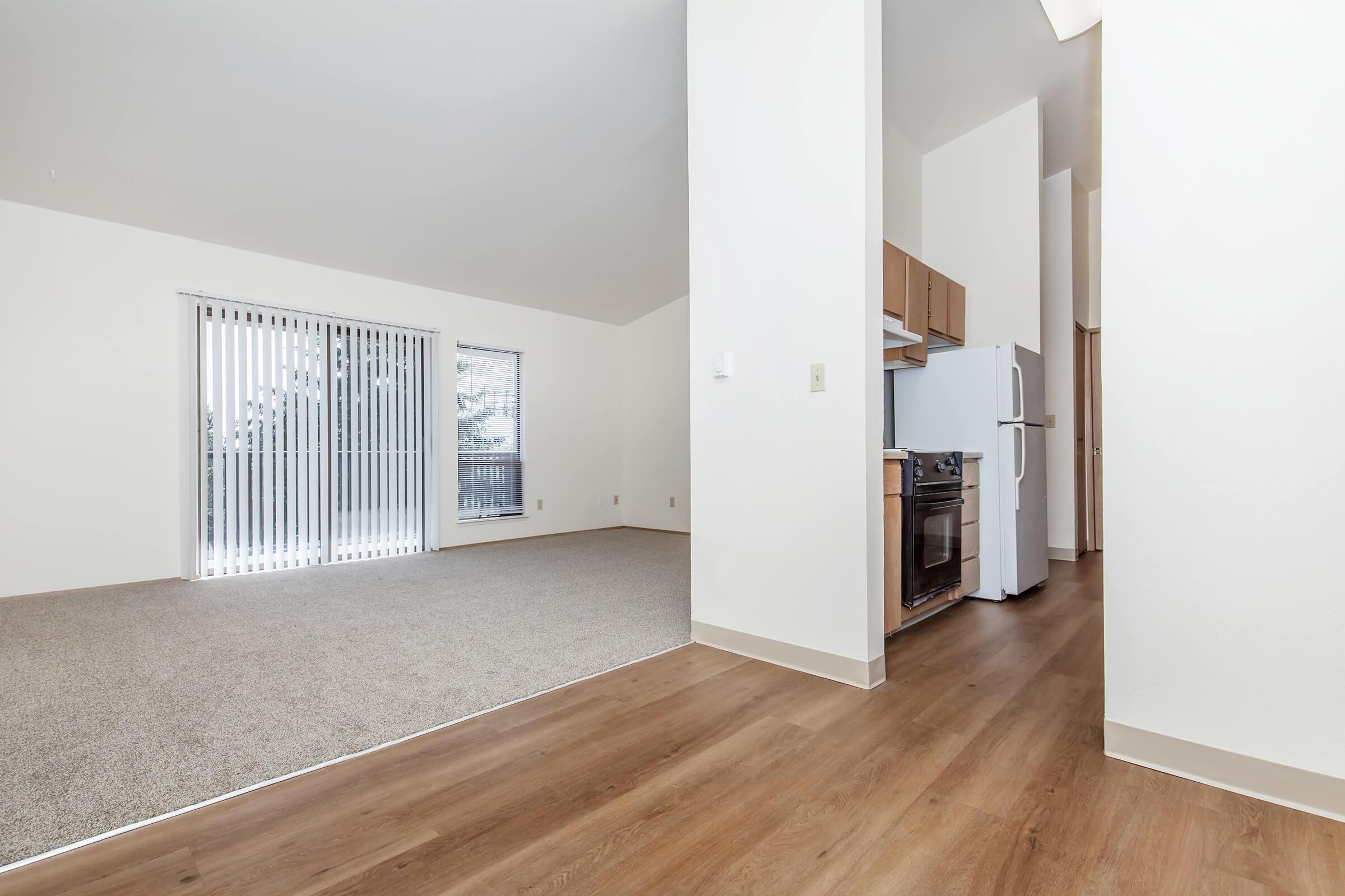 A spacious interior of an apartment featuring light-colored walls, carpet flooring, and vinyl plank flooring. On the left, there are sliding glass doors with vertical blinds, leading to a balcony. A kitchen area is visible in the background with an oven and refrigerator.