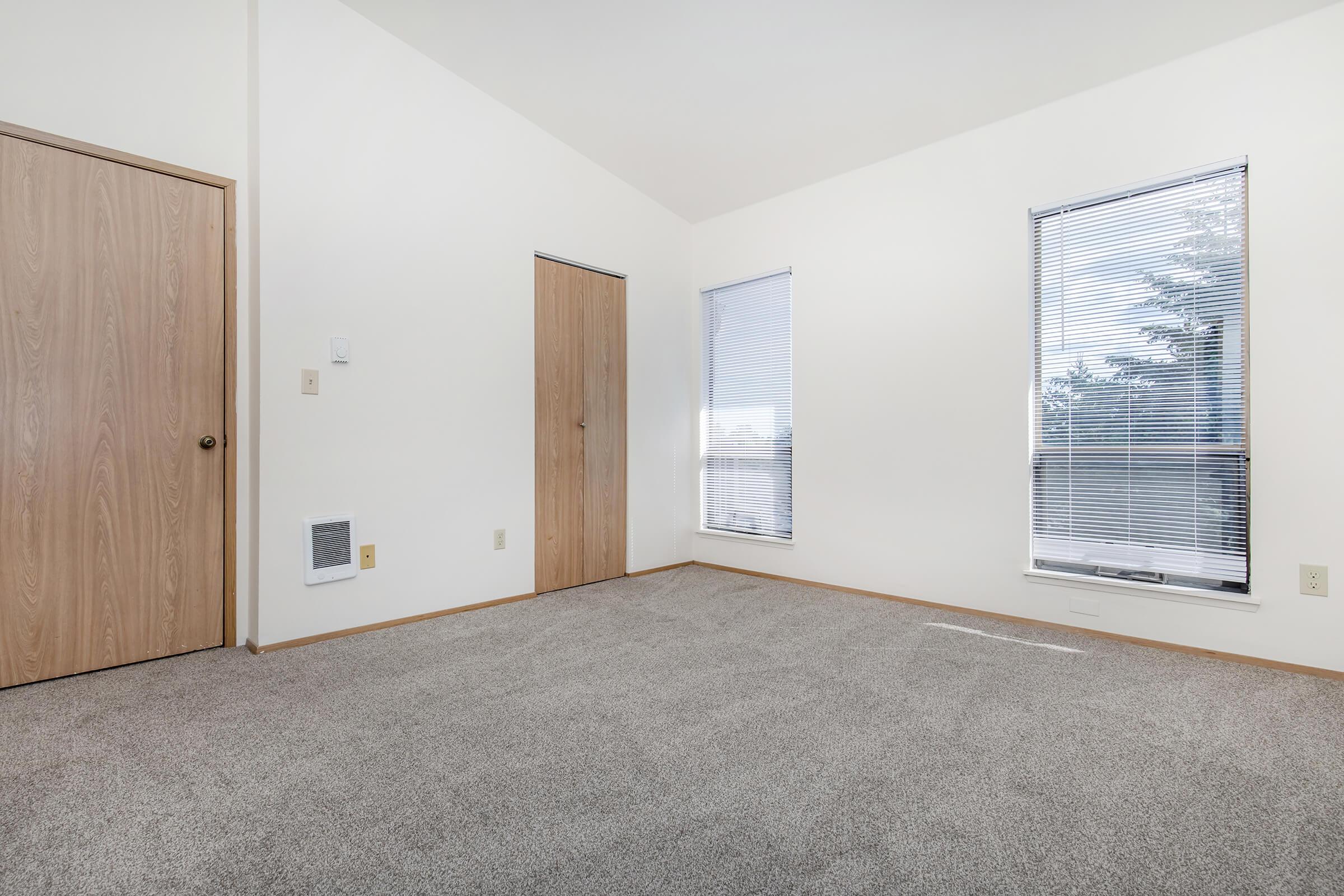 A bright, empty room with sloped ceilings, featuring a door on the left and a window on the right. The walls are painted white, and the floor is covered with light gray carpet. Natural light streams in through the window, creating an open and airy feel. A vent is visible on the wall near the floor.