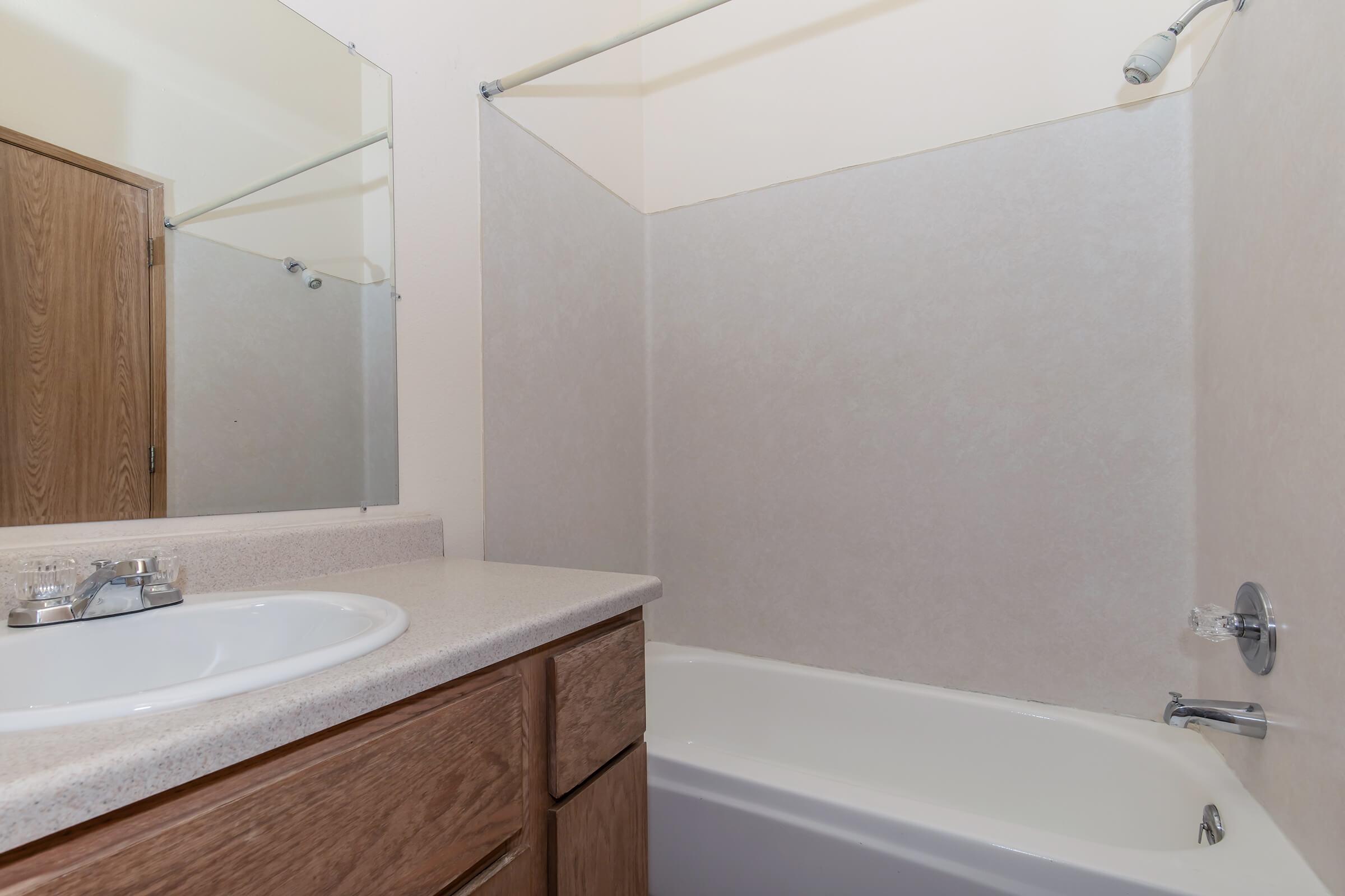 A clean, modern bathroom featuring a sink with a countertop, a wooden cabinet, a bathtub with a showerhead, and a large mirror. The walls are light-colored, and there is a simple design with no visible decorations. The overall atmosphere is tidy and inviting.