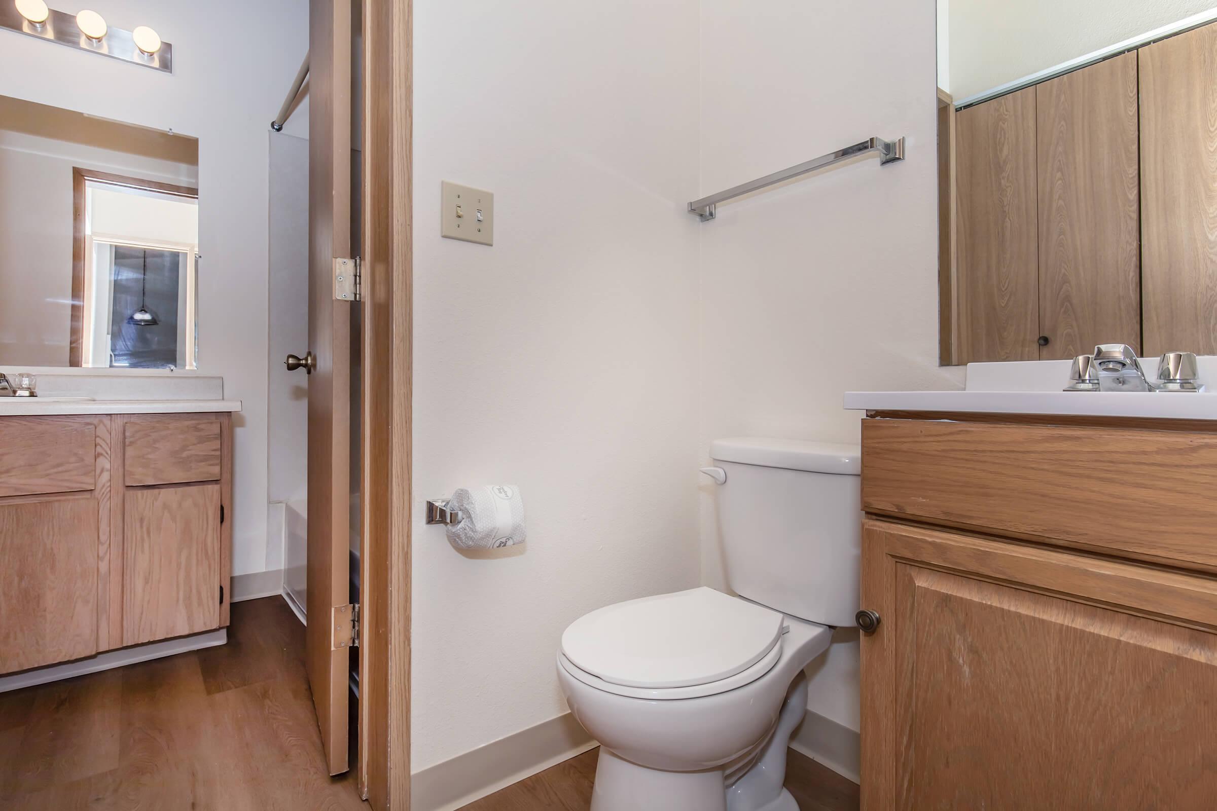 A clean and well-lit bathroom featuring a toilet, a sink with a mirror, and wooden cabinetry. The floor is finished with wood-like material, and a towel rack is mounted on the wall. A window allows natural light into the space, enhancing its overall brightness.