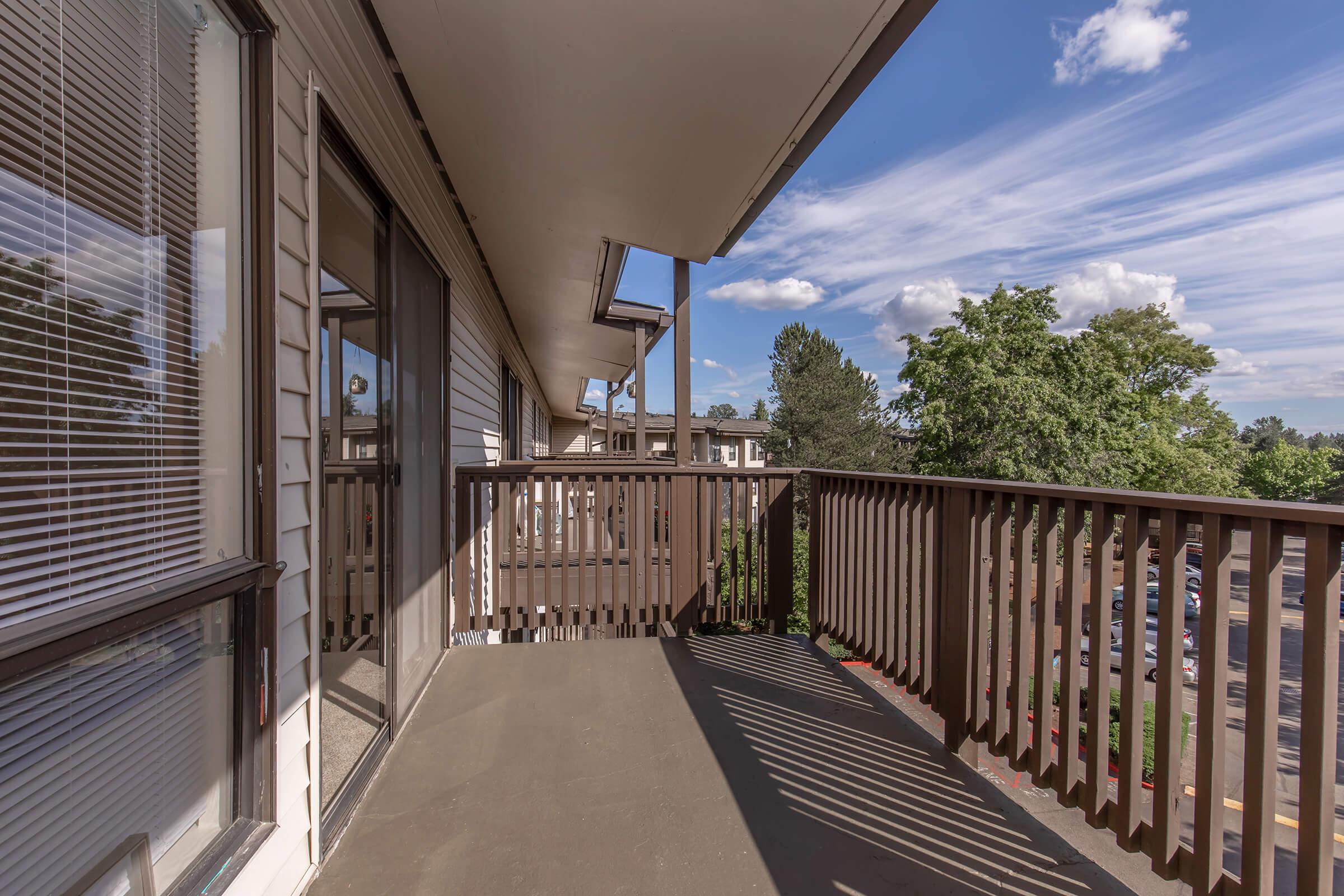 A spacious balcony with wooden railings overlooks a green area and parking lot. Bright, blue sky and clouds add a pleasant ambiance, while the sunlight creates shadows on the balcony floor. The scene is part of a multi-unit residential building, showing an inviting outdoor space.