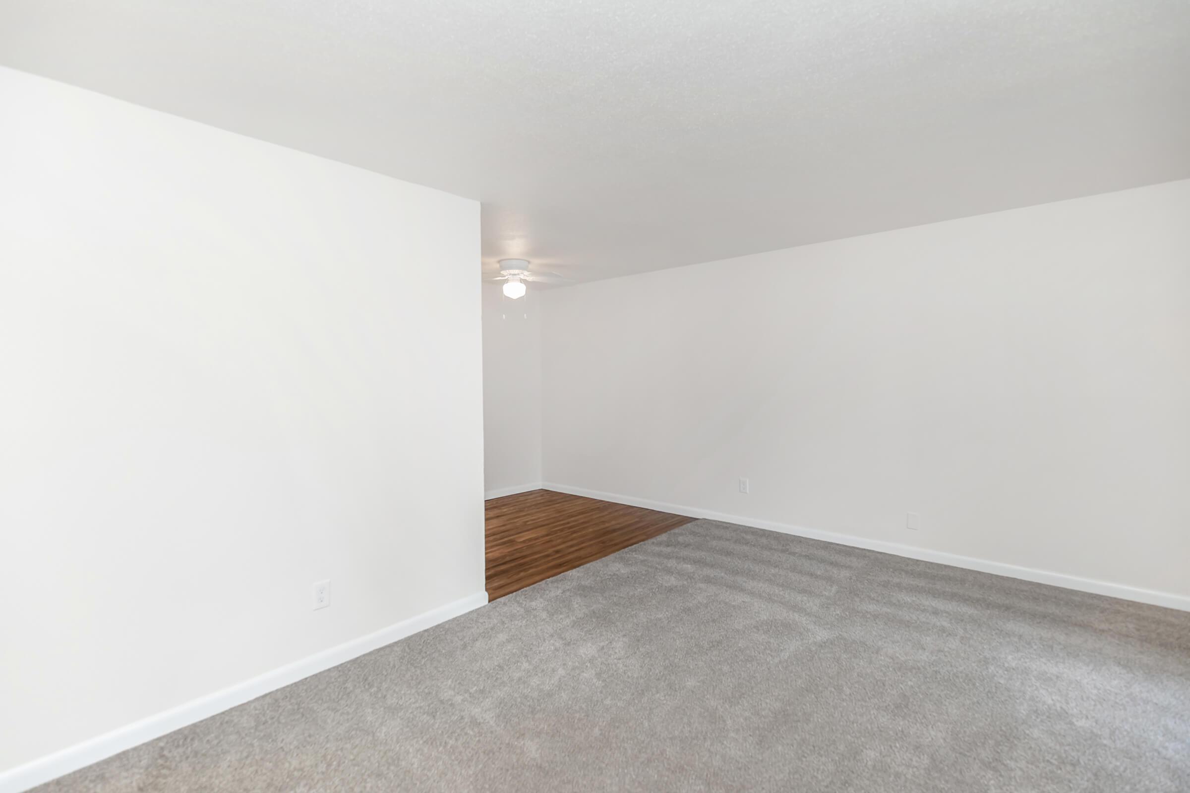 A spacious, empty room with light gray carpet, white walls, and a ceiling fan. There is a section with hardwood flooring visible in the background, creating a contrast with the carpeted area. The lighting is bright, emphasizing the openness of the space.