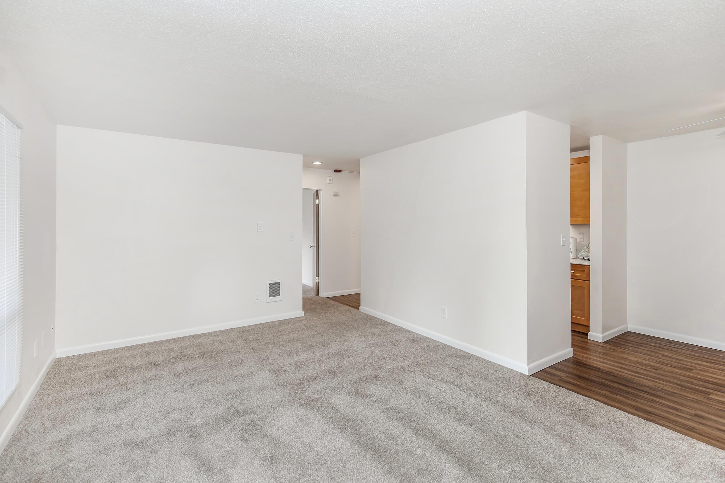 A spacious, empty living area with light-colored carpet and white walls. A doorway leads to another room in the background, and a small kitchen area is partially visible to the right. The space is bright and open, providing a welcoming atmosphere.