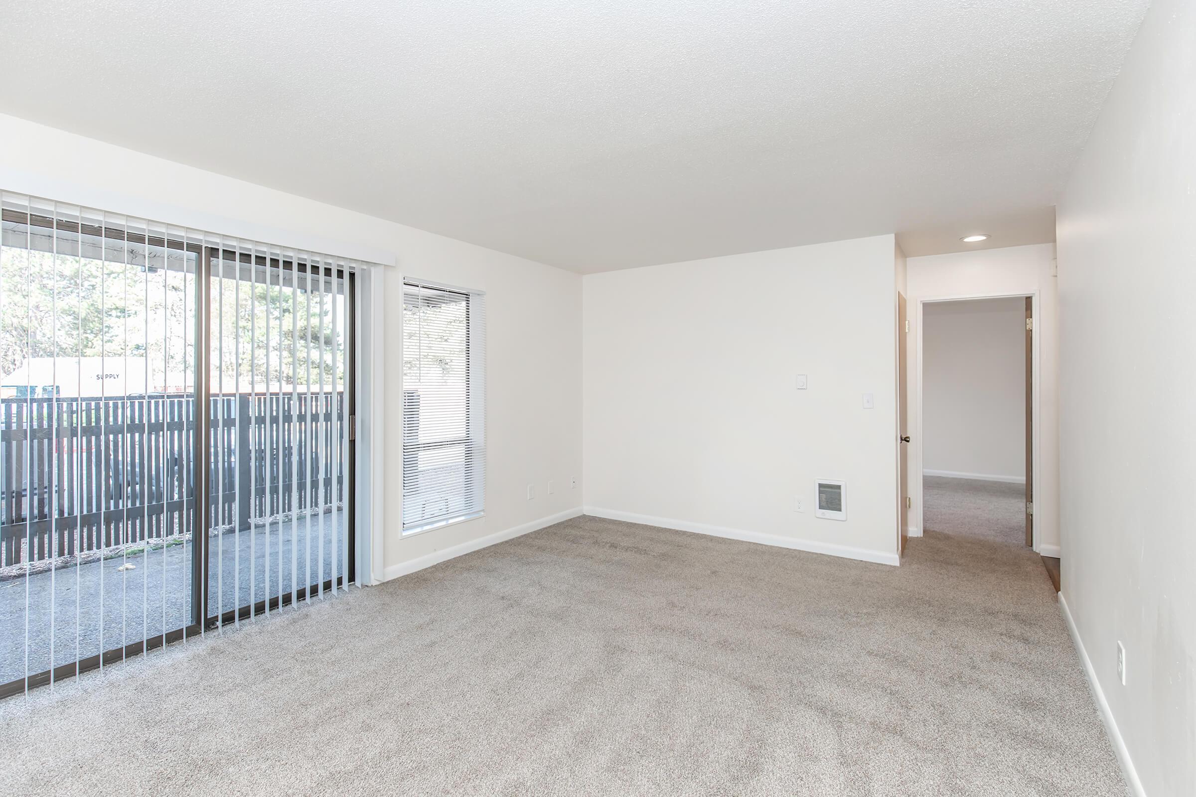 A spacious, unfurnished living room with light-colored walls and soft carpet. Large windows with blinds offer natural light and a view of an outdoor patio. A hallway leads to a door, suggesting access to other rooms or areas.