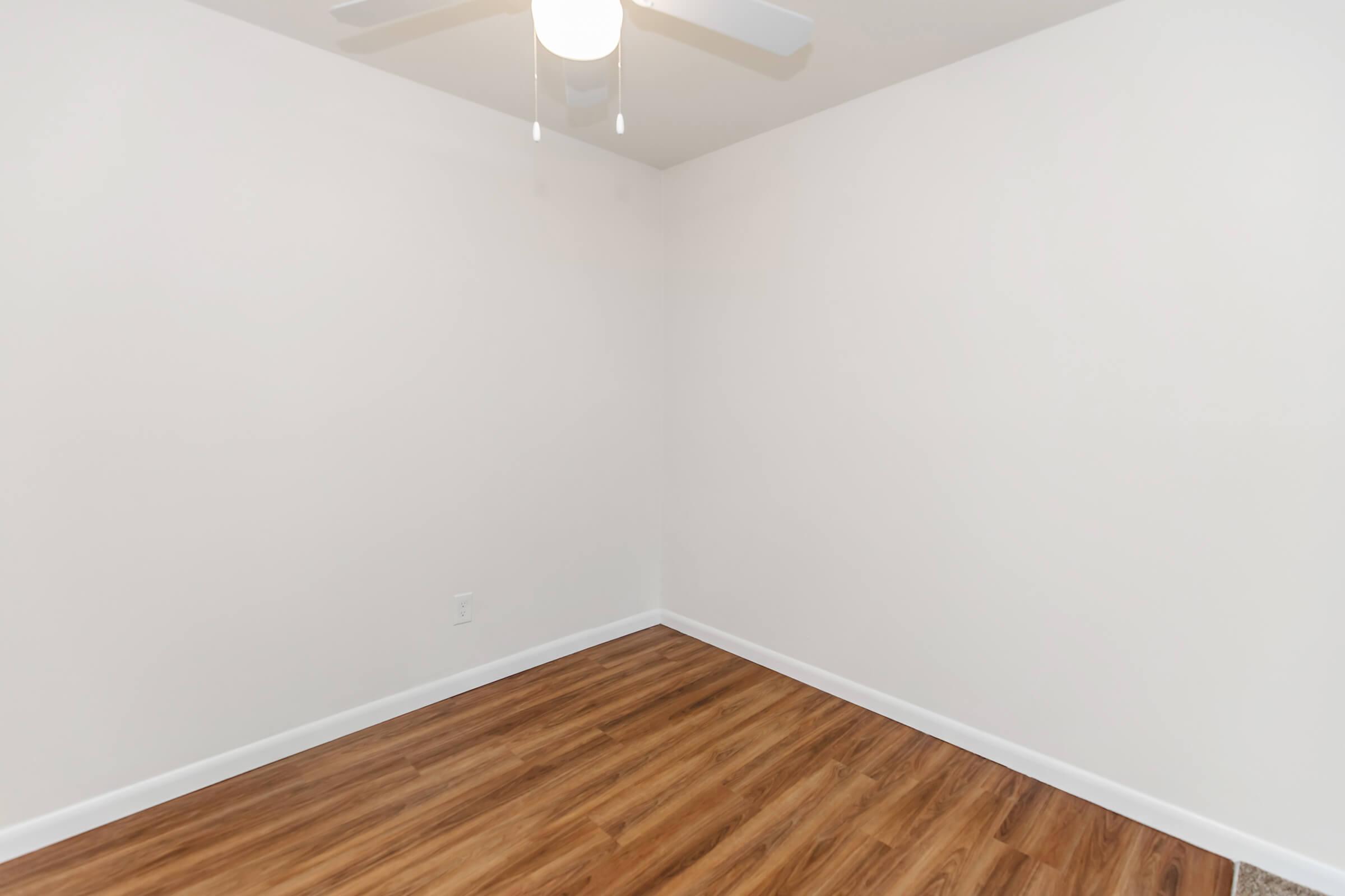A clean, empty room with light-colored walls and a wooden laminate floor. A ceiling fan hangs from the ceiling, providing minimal decor. The space is bright and airy, ideal for personalization or as a blank canvas for furnishings.