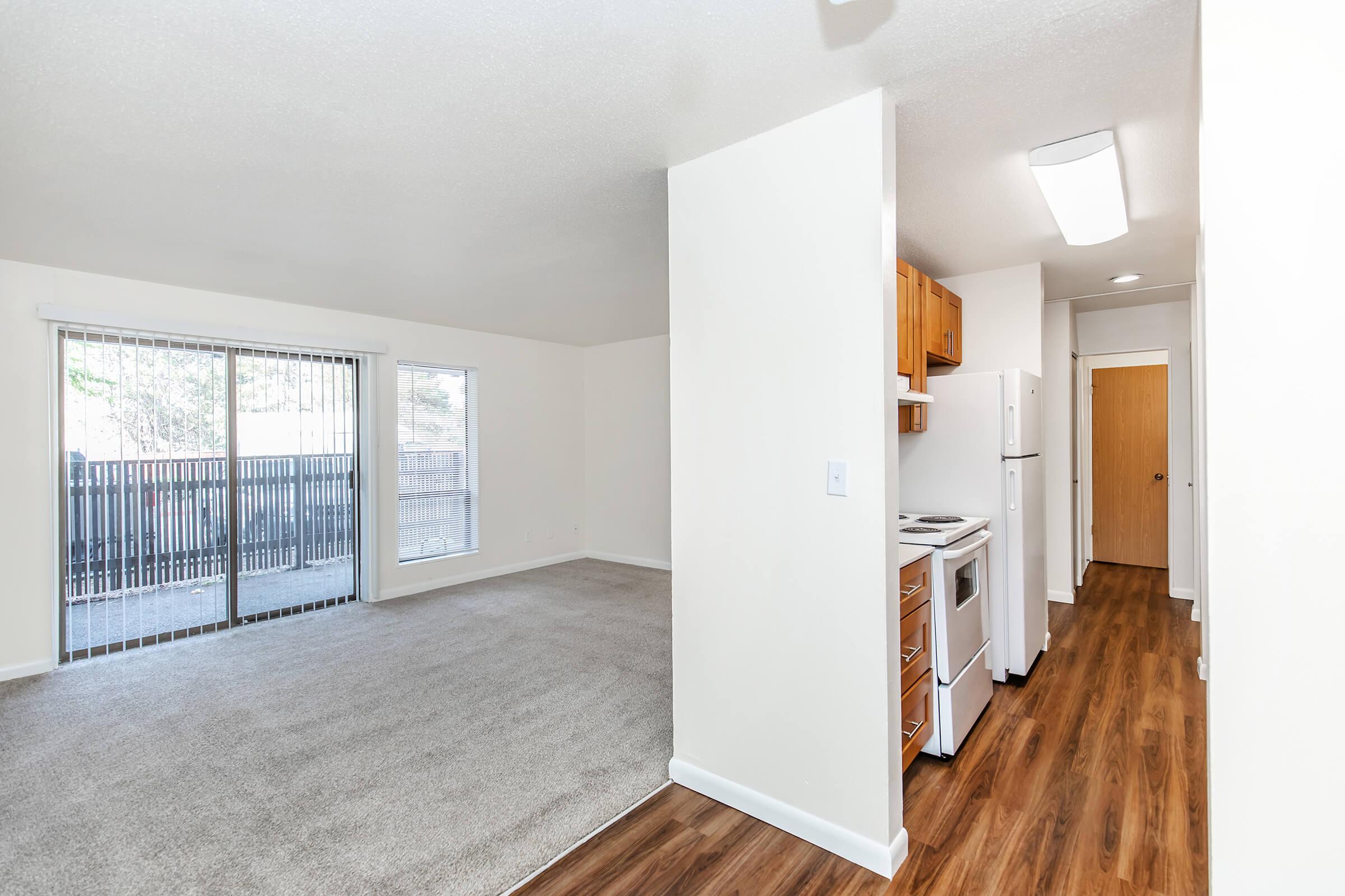 A spacious, brightly lit apartment interior featuring a living area with carpeted flooring, a nearby kitchen with modern appliances, and large sliding glass doors leading to an outdoor patio. Neutral-colored walls and warm wood accents create a welcoming atmosphere.