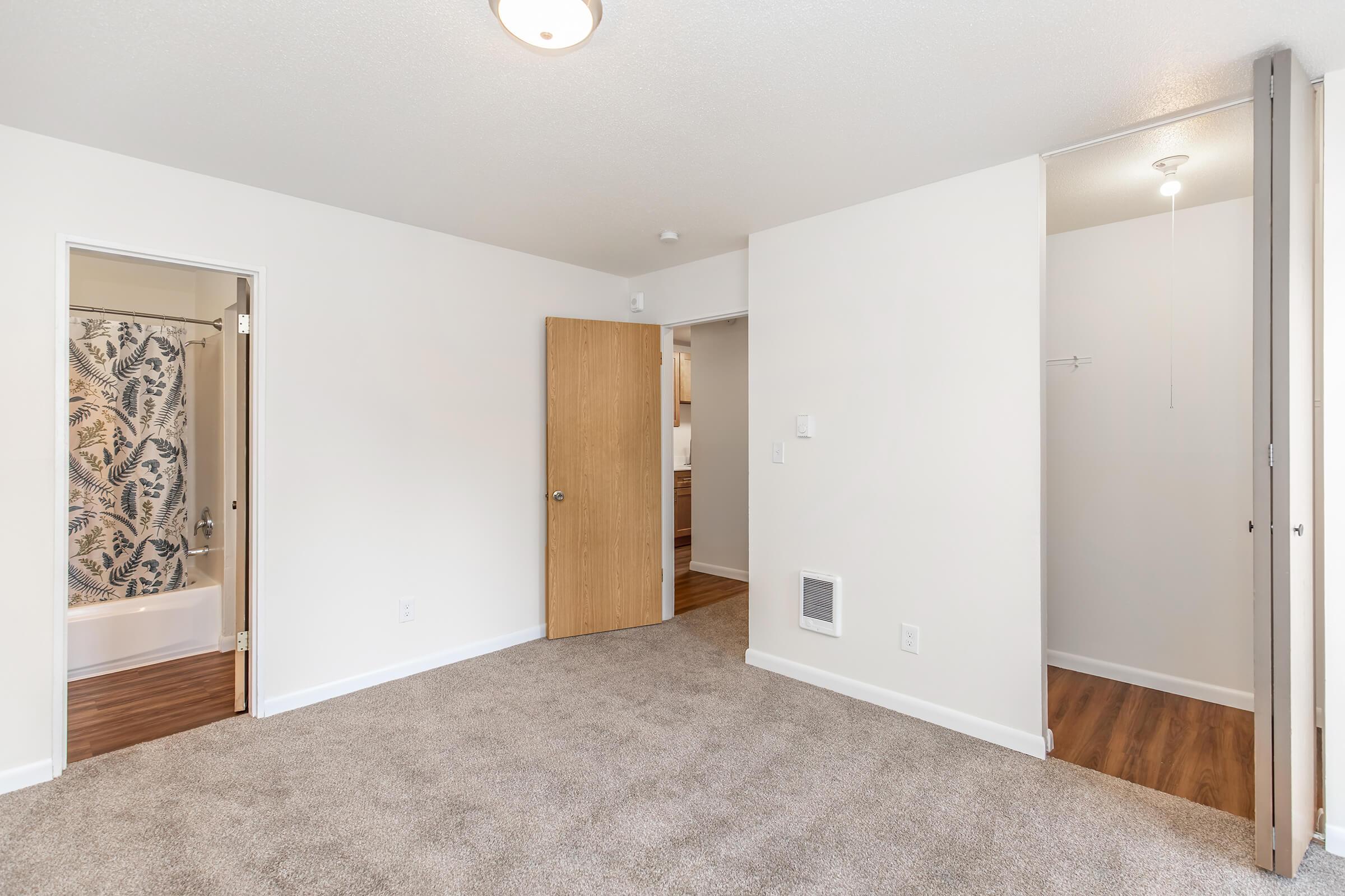 A bright, empty room with light-colored walls and plush carpet. Two doorways lead to other spaces: one opens to a bathroom with a shower curtain, while the other leads to a closet. There is a ceiling light fixture above, and the flooring near the entrances features hardwood.