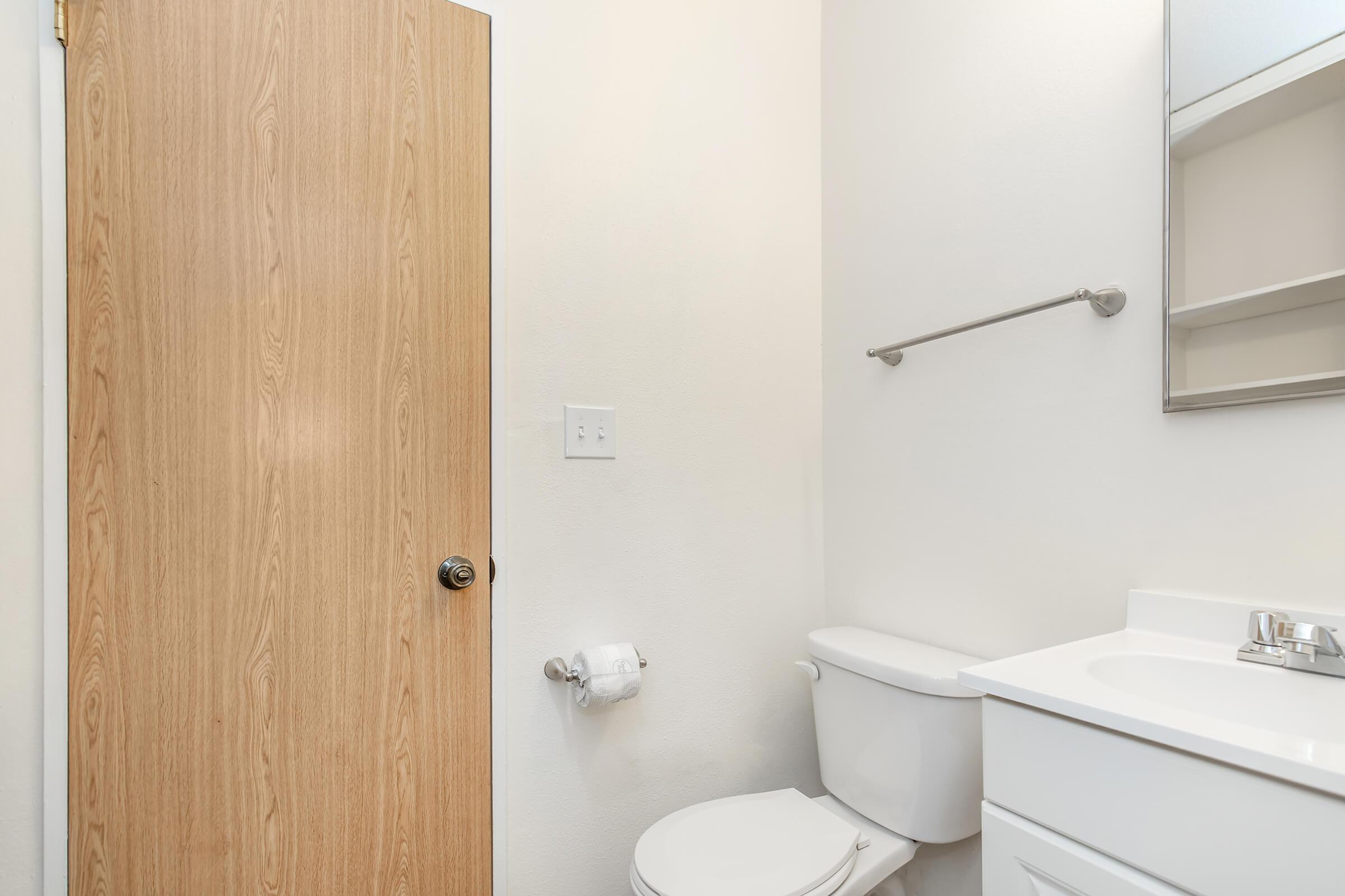 A simple bathroom interior featuring a light-colored wall, a wooden door, a white toilet, a small sink with a faucet, and a towel bar. There is also a toilet paper holder and a mirror above the sink. The overall design is clean and minimalistic.