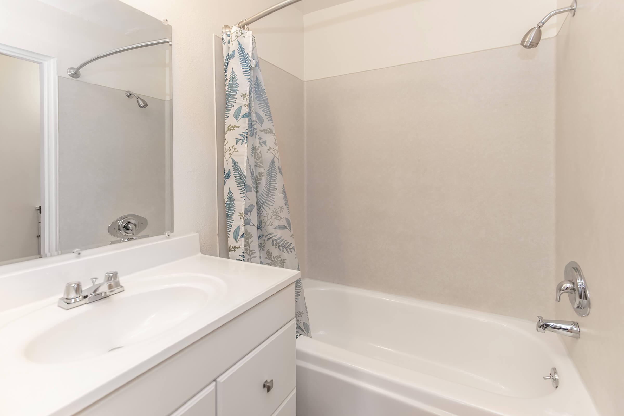 A clean bathroom featuring a white bathtub, a shower with a glass door, and a mirror above a sink. The bathroom walls are light-colored, and a shower curtain with a floral pattern hangs from a silver rod. The space is well-lit and tidy, emphasizing a minimalist design.
