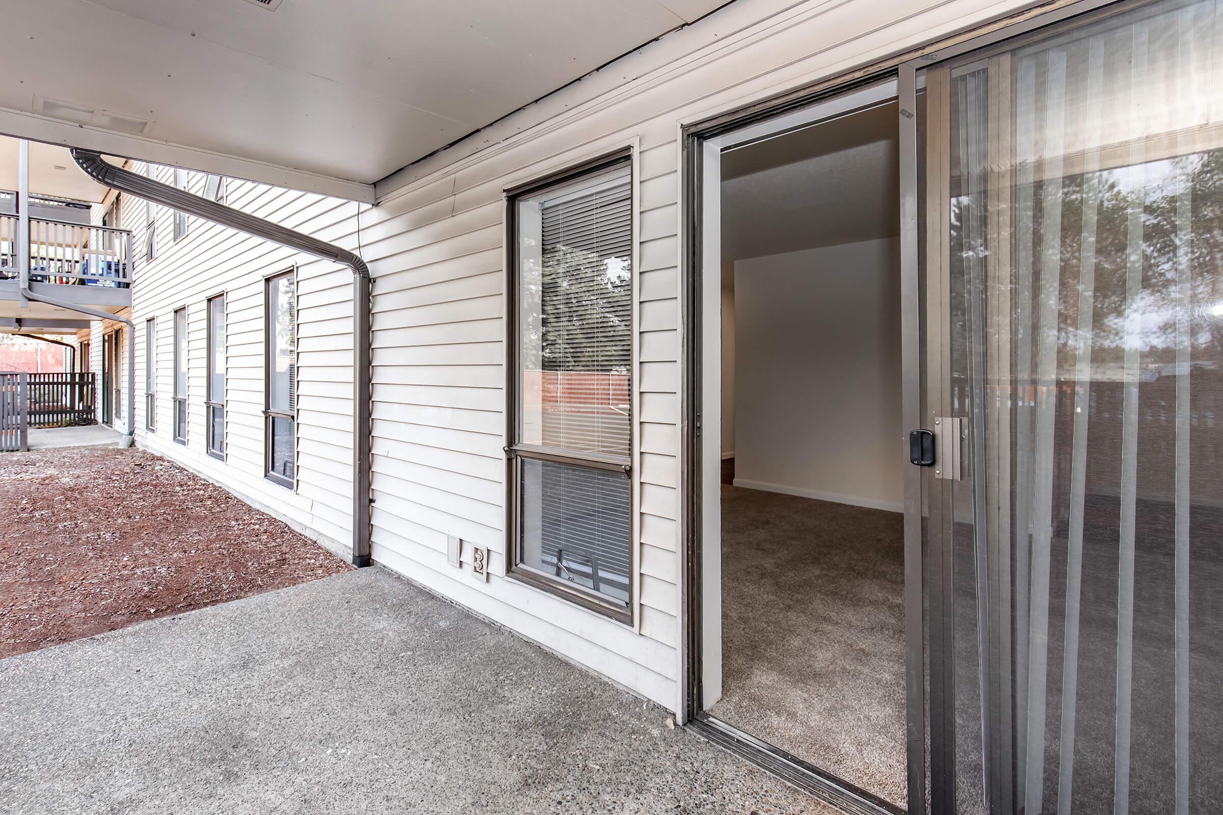 View of a patio area outside an apartment. The entrance features a sliding glass door leading into a carpeted interior space. The building has several windows visible along the exterior wall, with a gravel ground surface and an accessible entryway.