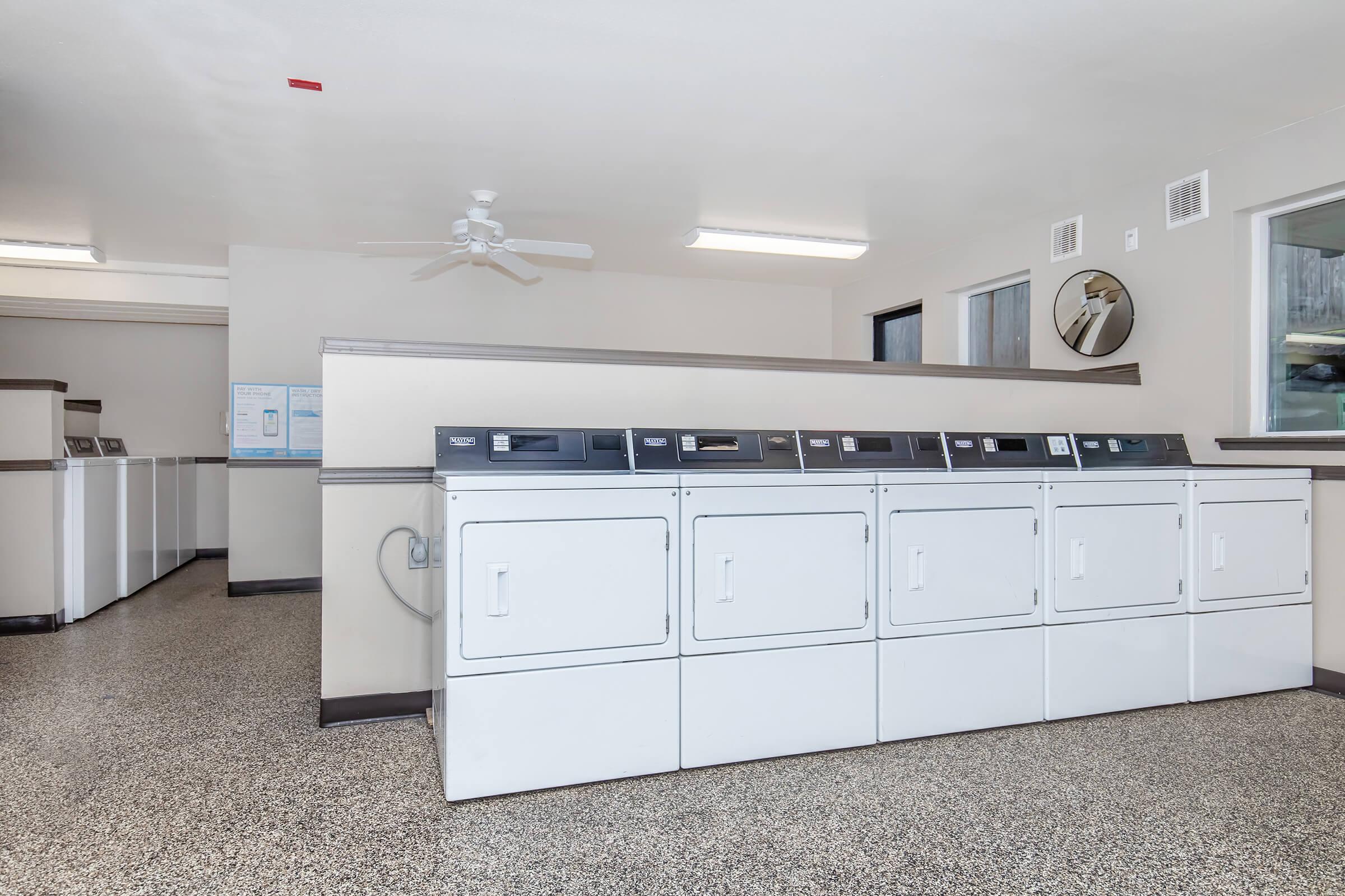 A laundry room featuring several white dryers and washing machines lined up against the wall, with a ceiling fan overhead. Natural light enters from windows, and there is a round mirror on the wall. The floor has a speckled design, contributing to a clean, functional environment for laundry tasks.