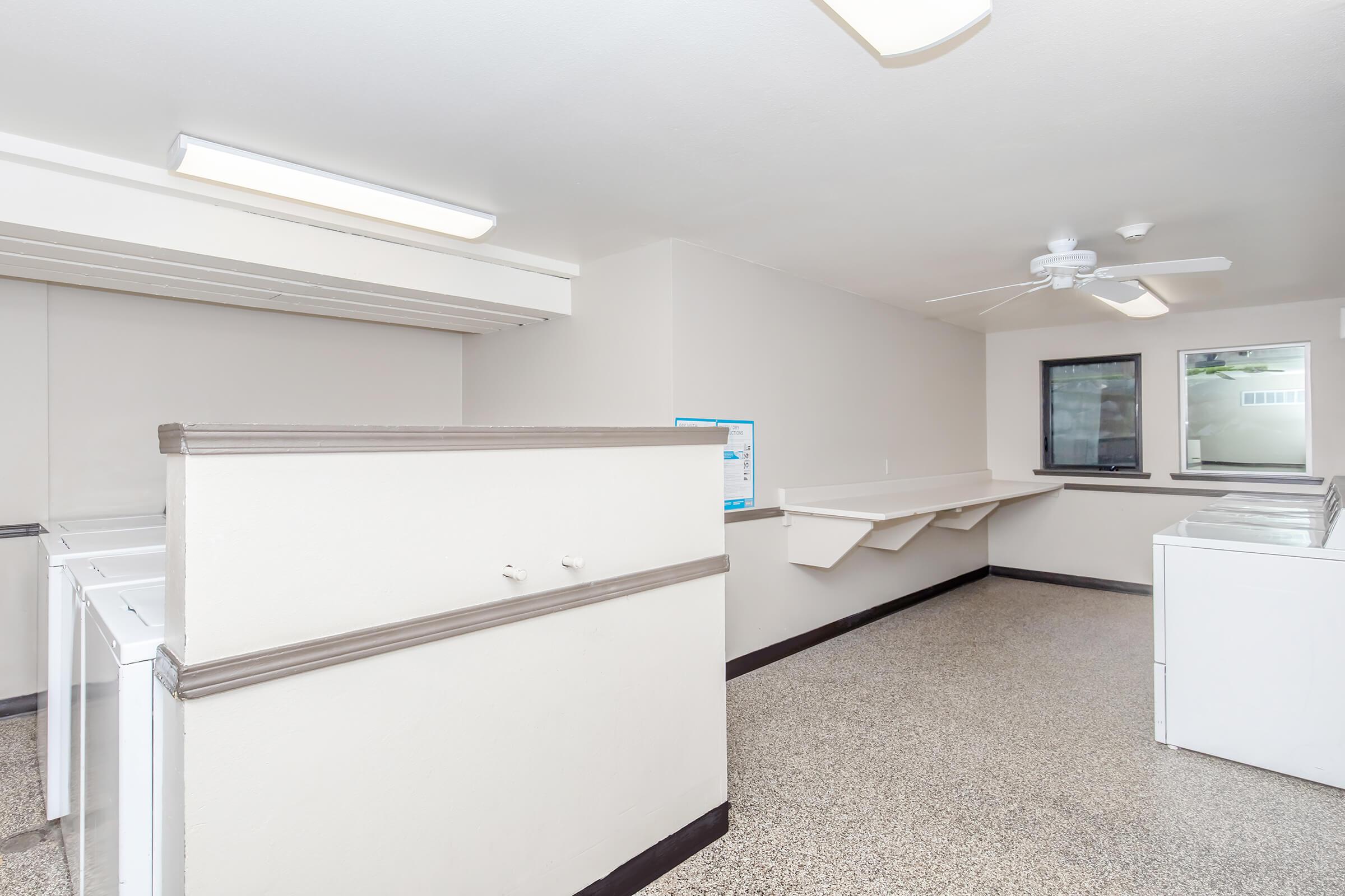 A spacious laundry room featuring several white washing machines and dryers. The room has a light-colored wall, overhead fluorescent lighting, and a ceiling fan. There are shelves along the wall and a window providing natural light, creating a bright and clean environment for laundry tasks.
