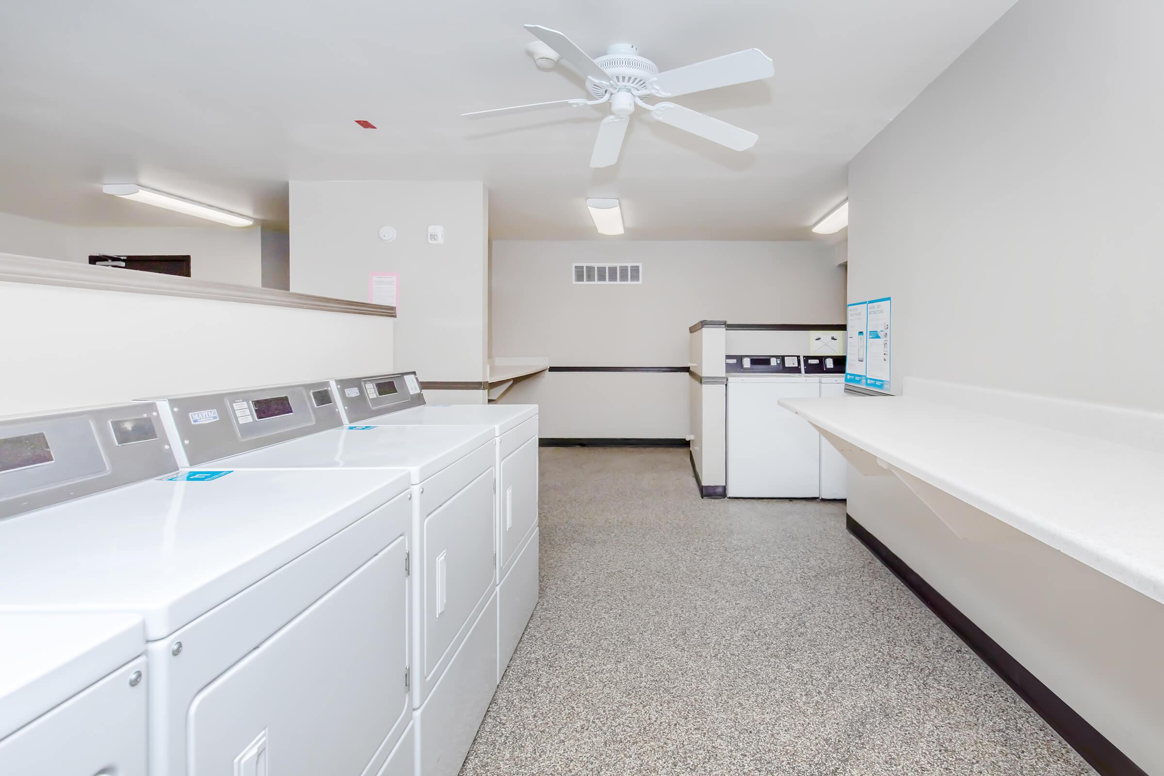 Bright and spacious laundry room featuring multiple white washing machines and dryers lined up against the wall. A ceiling fan is visible, providing ventilation. The room has light-colored walls and a clean, modern aesthetic with countertops for folding laundry. Natural light may illuminate the space.