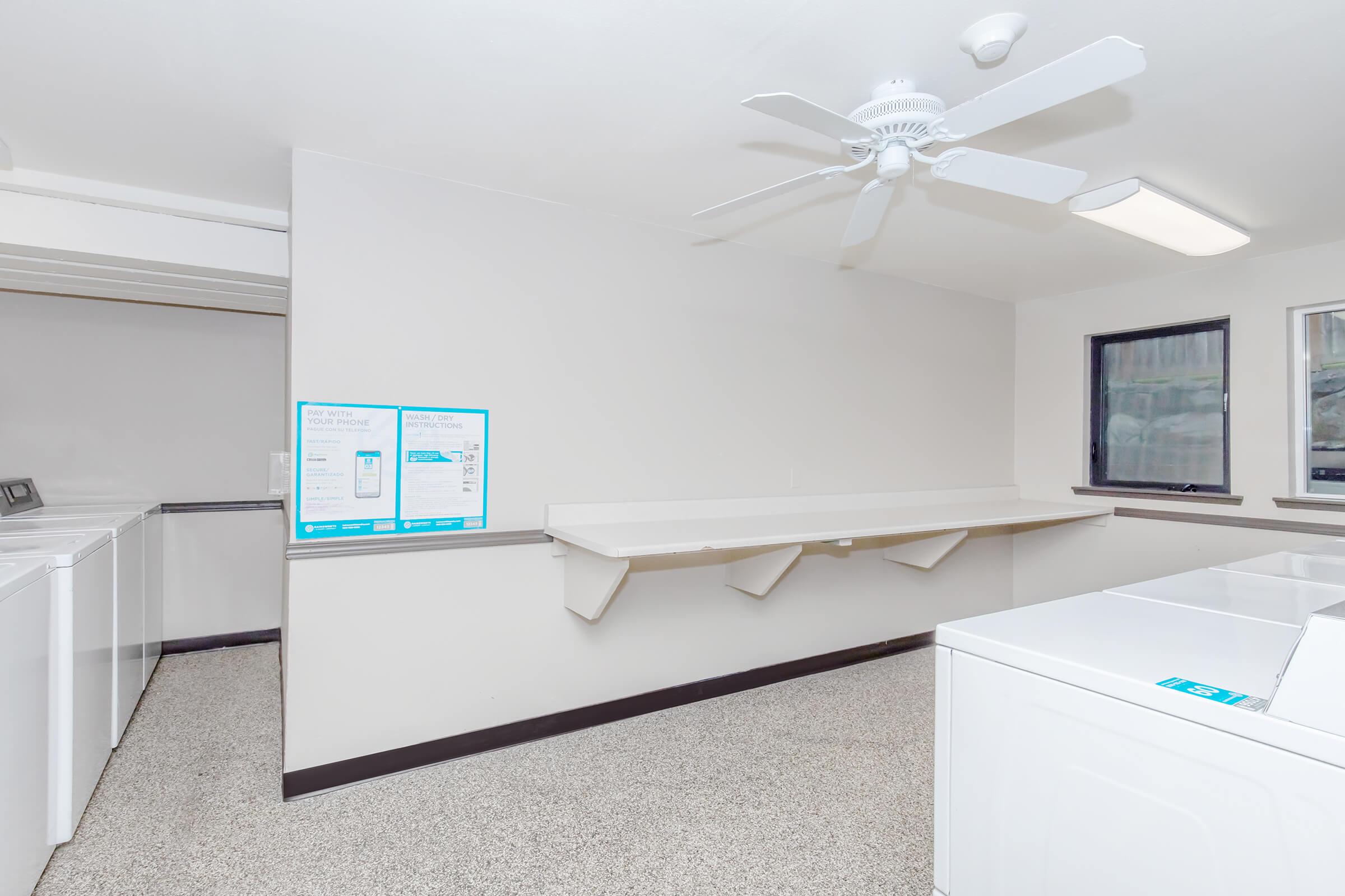 A clean and spacious laundry room featuring several washing machines, a folding area with a long countertop, and a ceiling fan. The walls are light-colored, and there are windows allowing natural light in, creating a bright atmosphere. Informational posters are visible on the wall.