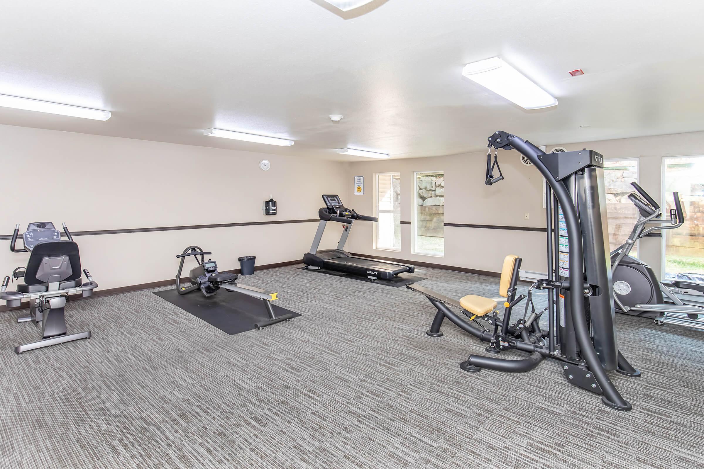 A bright and spacious gym area featuring exercise equipment including a treadmill, rowing machine, and multi-gym machine. The room has neutral-colored walls, ample lighting, and windows allowing natural light, with a clean and modern design.