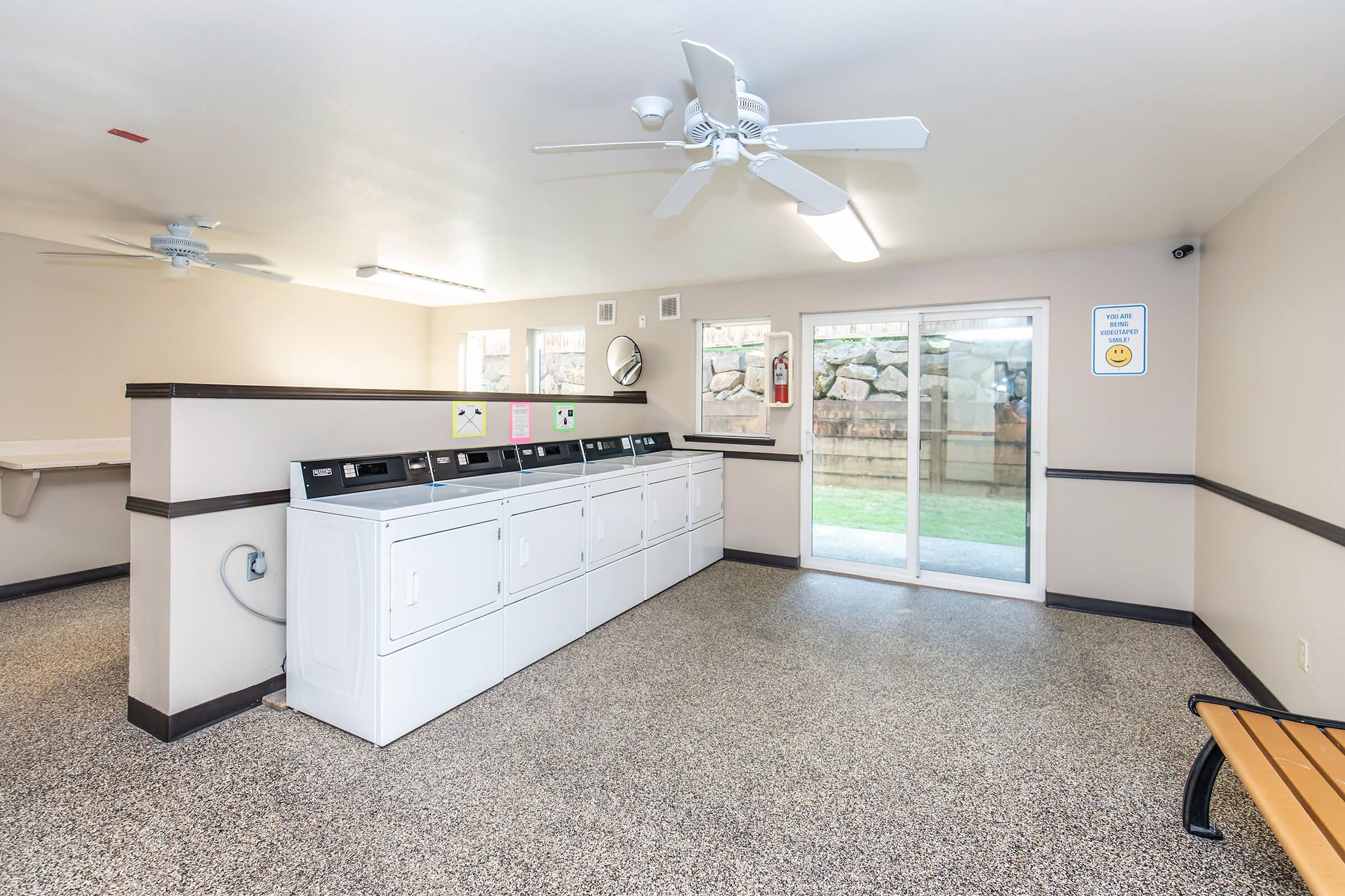 A clean and well-lit laundry room featuring multiple washing machines and dryers along one side, with a bench for seating. Large windows provide natural light, and there are signs for laundry instructions and safety. The space has a modern, inviting feel with a neutral color palette.