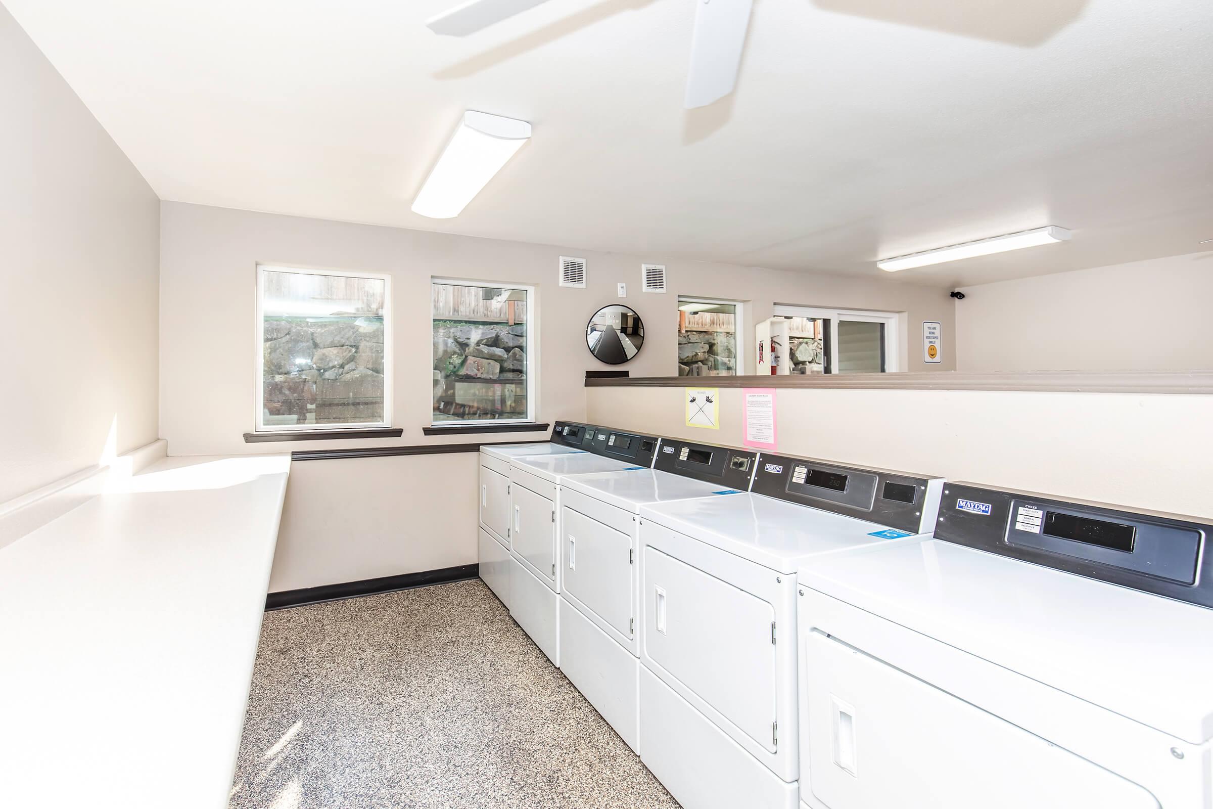 A bright and clean communal laundry room with multiple white washing machines and dryers. The walls are light-colored, with large windows allowing natural light to enter. A fan is mounted on the ceiling, and a countertop for folding laundry is visible against one wall. The flooring is textured.