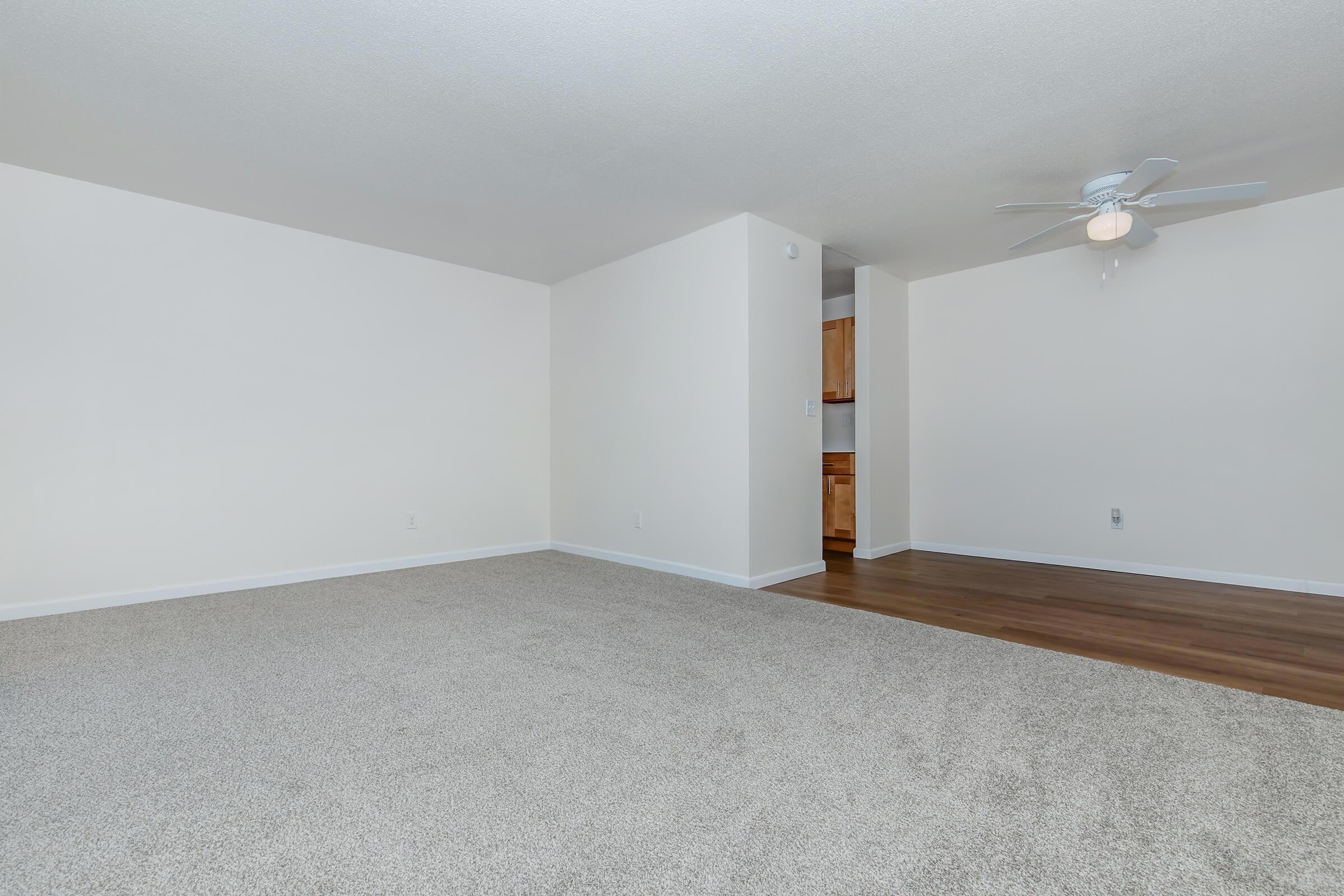 A spacious, empty room with light-colored walls and soft carpet flooring. A ceiling fan is visible, and a doorway leads to a kitchen area in the background. The overall atmosphere is bright and inviting, suitable for various furniture arrangements.