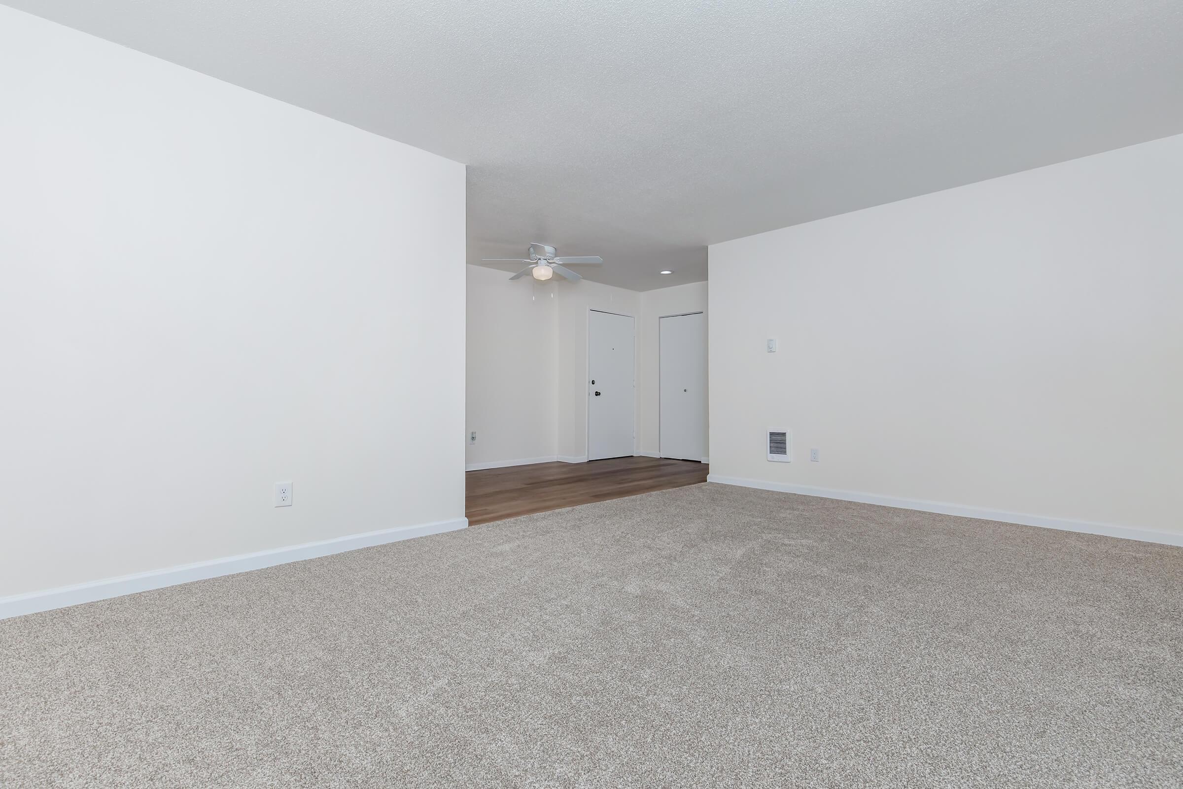 A spacious, empty living room featuring light beige carpet, a ceiling fan, and white walls. Two closed doors are visible in the background, leading to other rooms. The room is well-lit, creating a bright and open atmosphere.