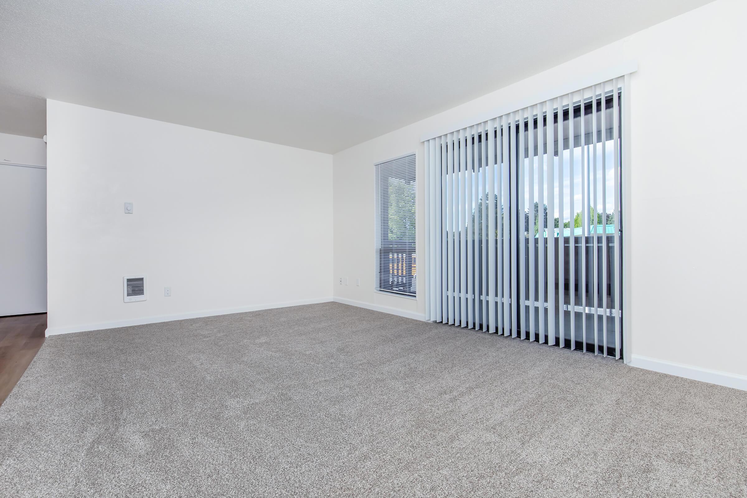 A spacious, empty living room featuring beige carpeted flooring and light-colored walls. Large sliding glass doors with white vertical blinds lead to a balcony, allowing natural light to enter. A small window is also present, providing additional light and a view outside.