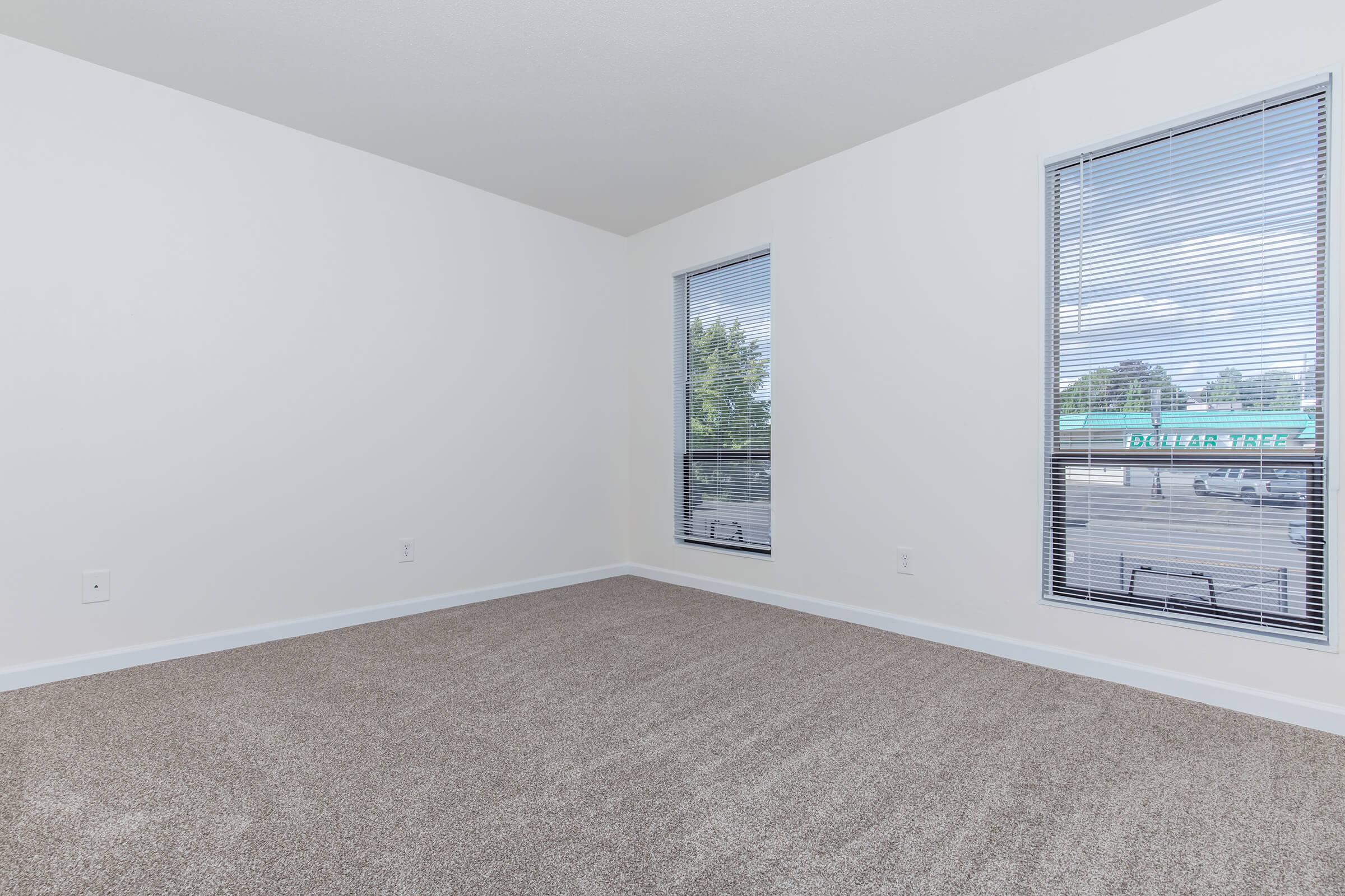 A vacant room with beige carpet flooring, two large windows with blinds allowing natural light, and white walls. The view through the windows shows a dollar store sign outside. The space appears bright and clean, suitable for various uses such as a living area or office.