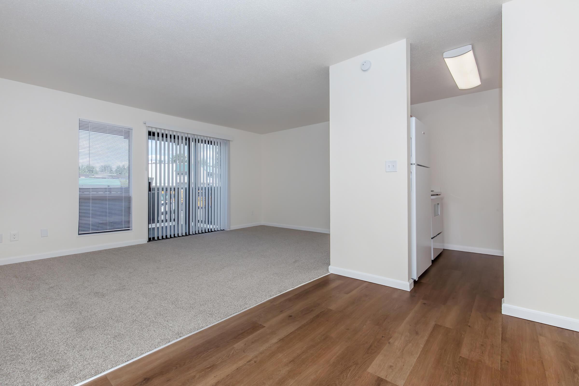 Spacious, well-lit apartment interior featuring a carpeted living area, large windows with vertical blinds, and a view towards a small balcony. The adjacent wall shows a kitchen area with a refrigerator. The floor has a mix of carpet and wood laminate, creating a cozy and modern atmosphere.